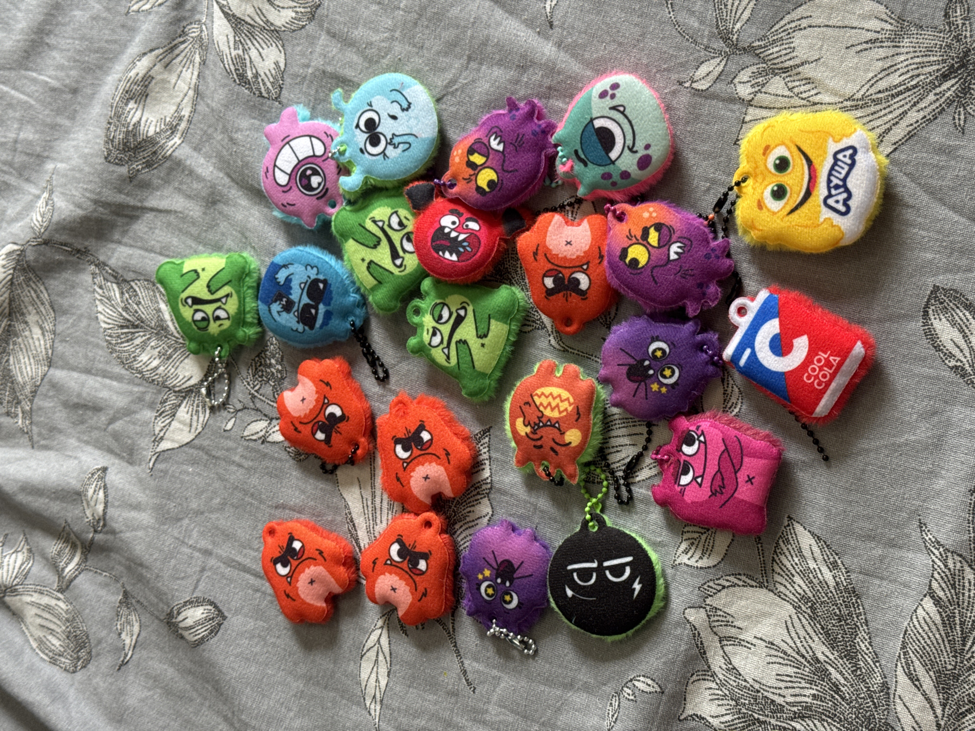 On a gray floral patterned surface are scattered 21 toy keychains with cartoon monster designs. The toy designs include monster faces, soda can shapes, and snack faces. The environment is devoid of any people or location context beyond the floral pattern background. The scene contains no indication of being taken in Chuprino, Russia.