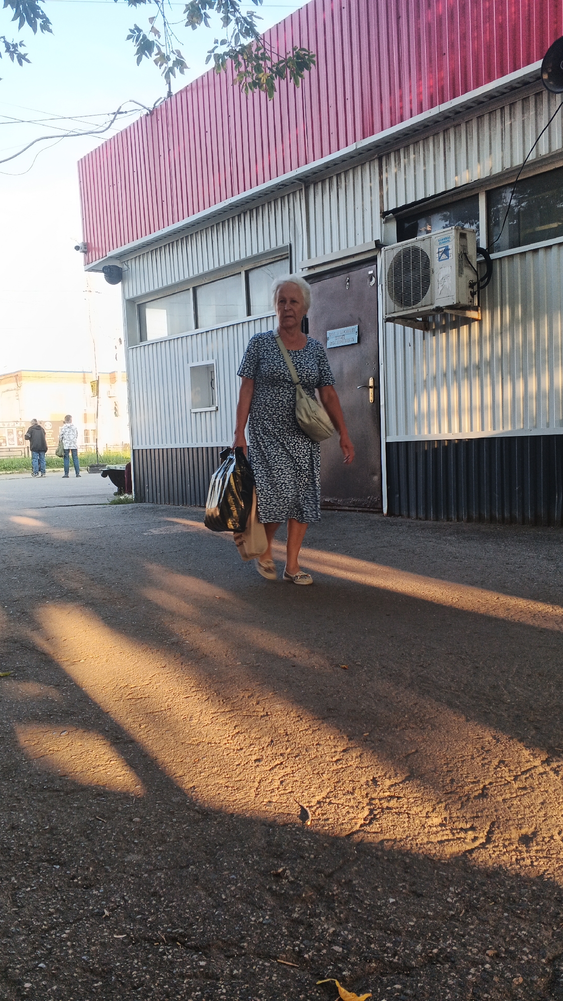 An elderly woman with short, light-colored hair is walking on an asphalt surface in front of a corrugated metal building with a red roof. She is wearing a dark blue dress with a white pattern and a light-colored crossbody bag. She is carrying two large black plastic bags in her right hand. The sun is casting long shadows across the ground. In the background, two figures are visible walking away from the camera, and a small black dog is sitting on the ground. There is an air conditioning unit mounted on the side of the building. The location is likely Izhevsk, Russia, based on the architecture and signage.