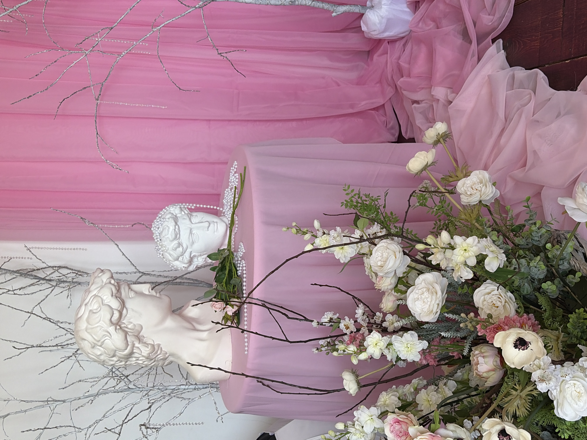 The scene presents a staged display, likely for photography or decorative purposes. Two white, classical-style busts are present, one with pearl ornamentation. The busts are positioned on and among pink fabric drapes. Floral arrangements in white and pale pink tones are placed around the busts and fabric. Painted white branches stand in the background. The environment suggests an indoor setup, possibly a studio or venue in Chaikovsky, Russia.