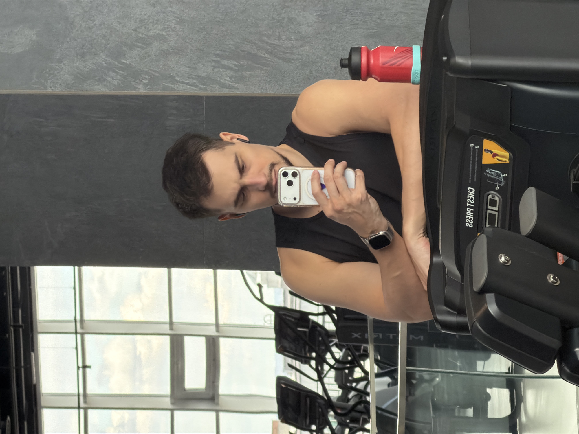 An image, originally rotated, depicts a man inside a modern gym in Krasnoyarsk, Russia. The man, with short dark hair, a goatee, and wearing a black sleeveless top, is holding a smartphone with a white case and a pop socket. He has an earbud in his left ear and a smartwatch on his left wrist, appearing to be taking a selfie or looking at his phone.
He is positioned next to a large black gym machine, likely a chest press, which occupies the right side of the frame. A red water bottle with a black cap and a light blue band is resting on this machine. In the background, a textured grey wall is visible behind the man, and large windows reveal an outdoor scene with bright daylight, suggesting it is daytime. The reflection of another gym equipment, bearing the brand "MATRIX," can be seen in the window.
The man's expression is neutral and focused on his phone. Visible text on the gym equipment reads "CHEST PRESS" (reversed as "22B ЧЭ") along with a diagram illustrating its use. The scene suggests a moment of rest or activity documentation during a workout session.