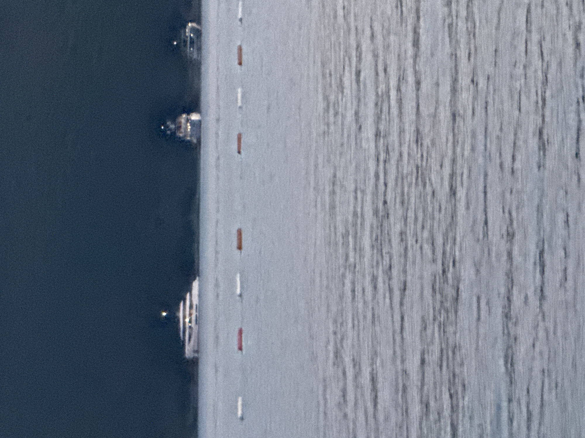 The image displays a body of water with several buoys floating on the surface. In the background, three yachts are visible, with the leftmost and largest yacht appearing to be a superyacht with multiple decks and illuminated windows. The middle and rightmost yachts are smaller. The water is calm, with gentle ripples reflecting the ambient light. The sky behind the yachts is dark and overcast. The scene is likely a harbor or bay in Boreti, Montenegro, given the presence of yachts and buoys. No people are discernible in the image.