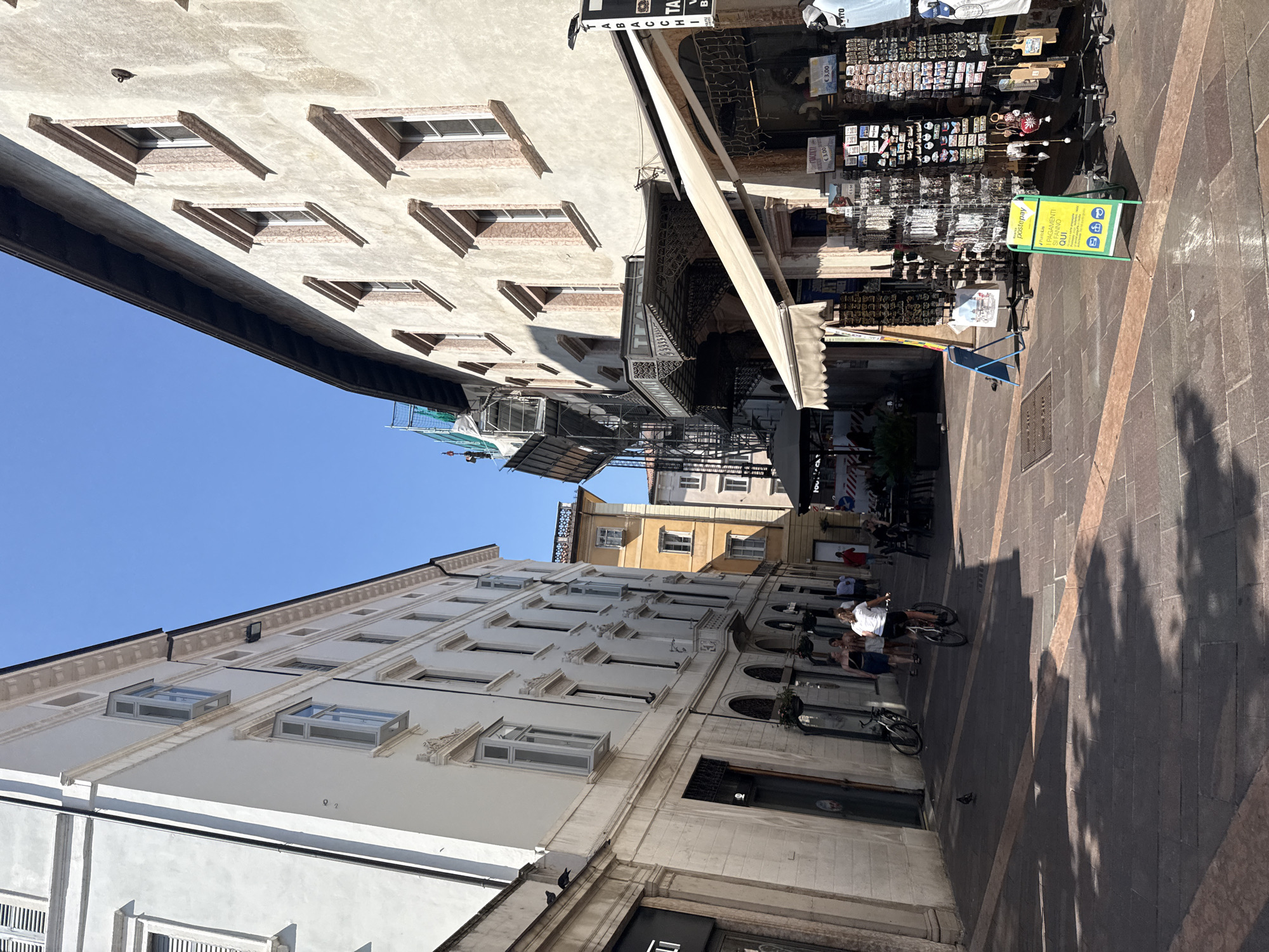 This image depicts a street scene in Trento, Italy, on a sunny day. The perspective looks down a narrow, cobblestone street flanked by tall buildings on both sides. The building on the left is painted white with numerous windows and a classical architectural style. The building on the right has a textured, off-white facade with decorative window frames.
In the foreground, a woman is riding a bicycle away from the camera. Several other people are walking or standing on the sidewalk. A small bird is visible on the ground. Along the right side of the street, there are shops with awnings and displays of merchandise, including souvenirs and keychains. A green sign with Italian text about payments is prominently displayed.
In the background, scaffolding is visible on the side of one of the buildings, suggesting ongoing construction or renovation. The sky is a clear, bright blue. The street is paved with dark cobblestones and features decorative linear patterns. Shadows from the buildings and possibly trees are cast across the pavement. A few bicycles are parked near the shops on the left. The overall atmosphere appears to be one of a bustling European city on a pleasant day.