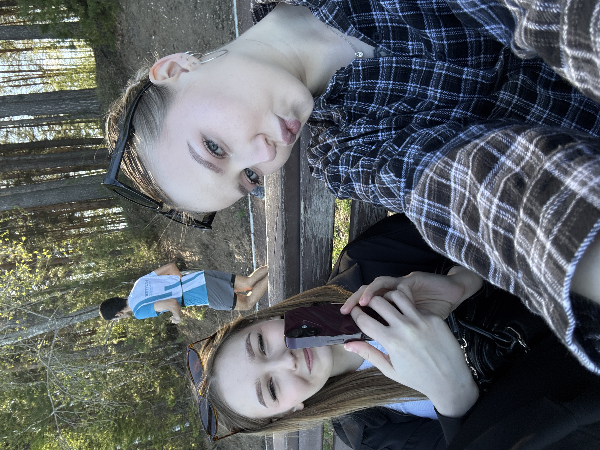 Two young women are seated on a bench, likely taking a selfie. The woman on top has blonde hair and is wearing a black and white plaid shirt. She has sunglasses perched on her head and is wearing jewelry. The woman below has brown hair and is holding a cell phone in front of her face. She is also wearing sunglasses on her head and a black jacket. In the background is a man wearing a blue shirt and gray shorts doing push-ups or stretching near trees. The environment appears to be a park or wooded area, likely in Chaikovsky, Russia.