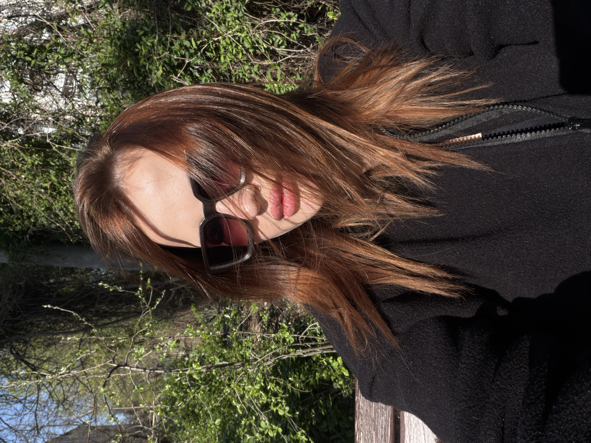 A woman with reddish-brown hair is seated outdoors on a wooden bench. She is wearing sunglasses and a black zippered jacket. The background features green vegetation, possibly trees and bushes. The location is not identifiable as Krasnodar, Russia based on the visual information provided.