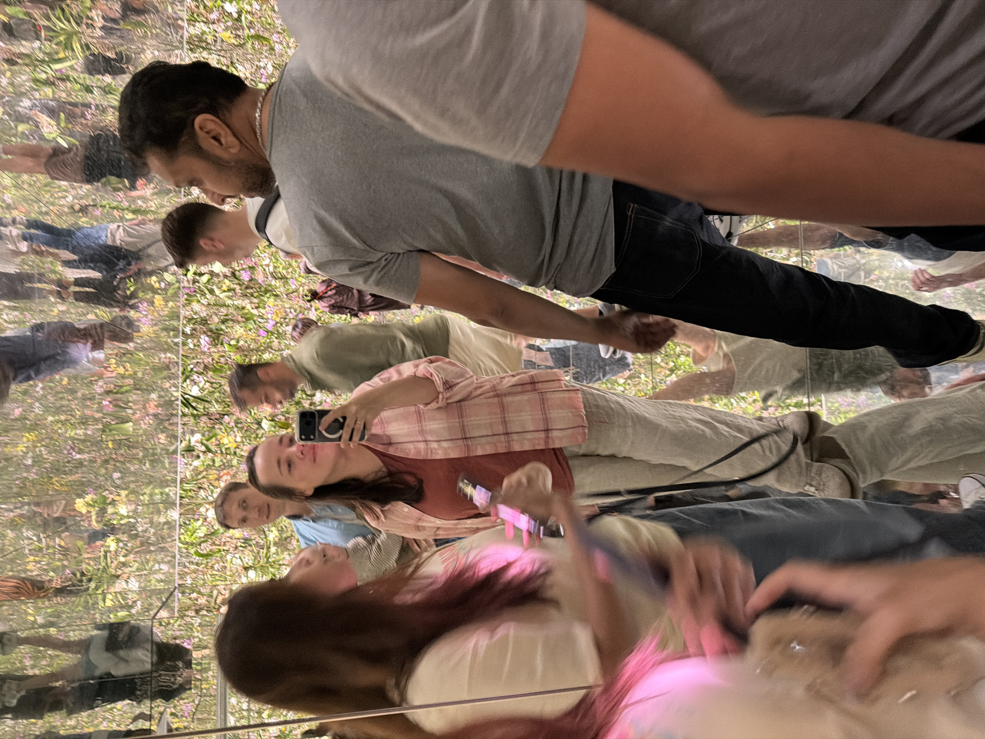 In Koto, Japan, this image depicts a group of individuals in a mirrored room with a floral, nature-inspired installation. The room's mirrored surfaces create a disorienting and immersive environment, reflecting the people and the installation from multiple angles, including upside down.
In the foreground, a woman in a pink plaid shirt and light-colored pants holds a smartphone, possibly taking a selfie. To her left, another person, partially obscured, wears a light-colored top and appears to be interacting with a phone. A man in a grey t-shirt and dark jeans stands in the mid-ground, facing right. Behind him, a man in a light blue shirt looks towards the woman with the phone, and another man in a green shirt is visible further back. The installation itself is a vibrant mix of green foliage and colorful flowers, artfully arranged to create a sense of being outdoors, despite the indoor setting. The overall impression is one of playful exploration and shared experience within an artistic space.