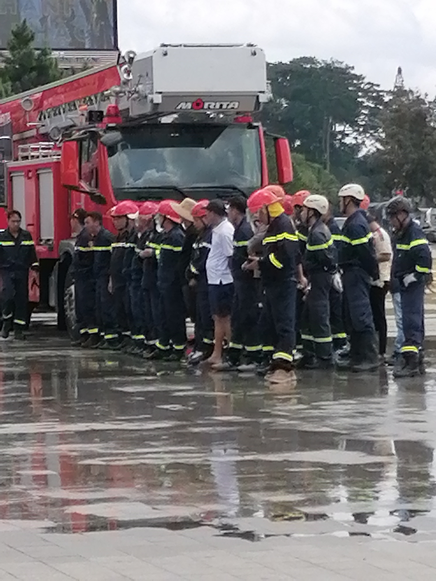 A group of firefighters in dark blue uniforms with yellow reflective stripes stand in a line, facing forward, on a wet, reflective surface, possibly pavement after rain. They are all wearing helmets, some red, some white. A large red fire truck with a ladder mechanism is parked behind them, with the "MORITA" brand visible on its upper section. The background includes trees and a cloudy sky. This scene is located in Da Lat, Vietnam, as indicated by the specific location in the city.