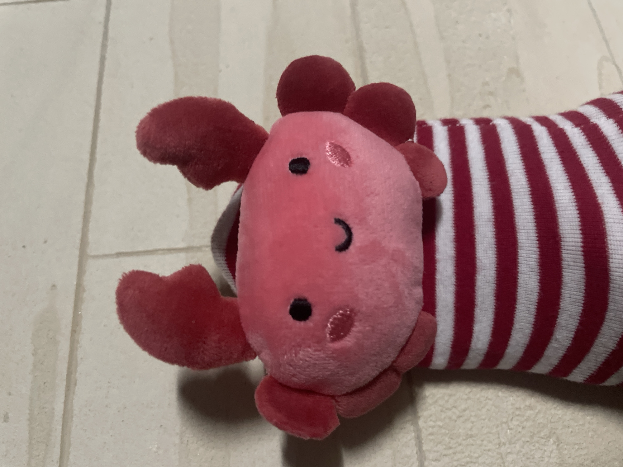 A plush toy, resembling a red crab with a pink head and a body covered in red and white stripes, lies on a surface with a light beige color. The toy has large claws and small spherical protrusions on its head. The surface has a slight texture. There is no visible information to suggest the location is Novi Sad, Serbia.