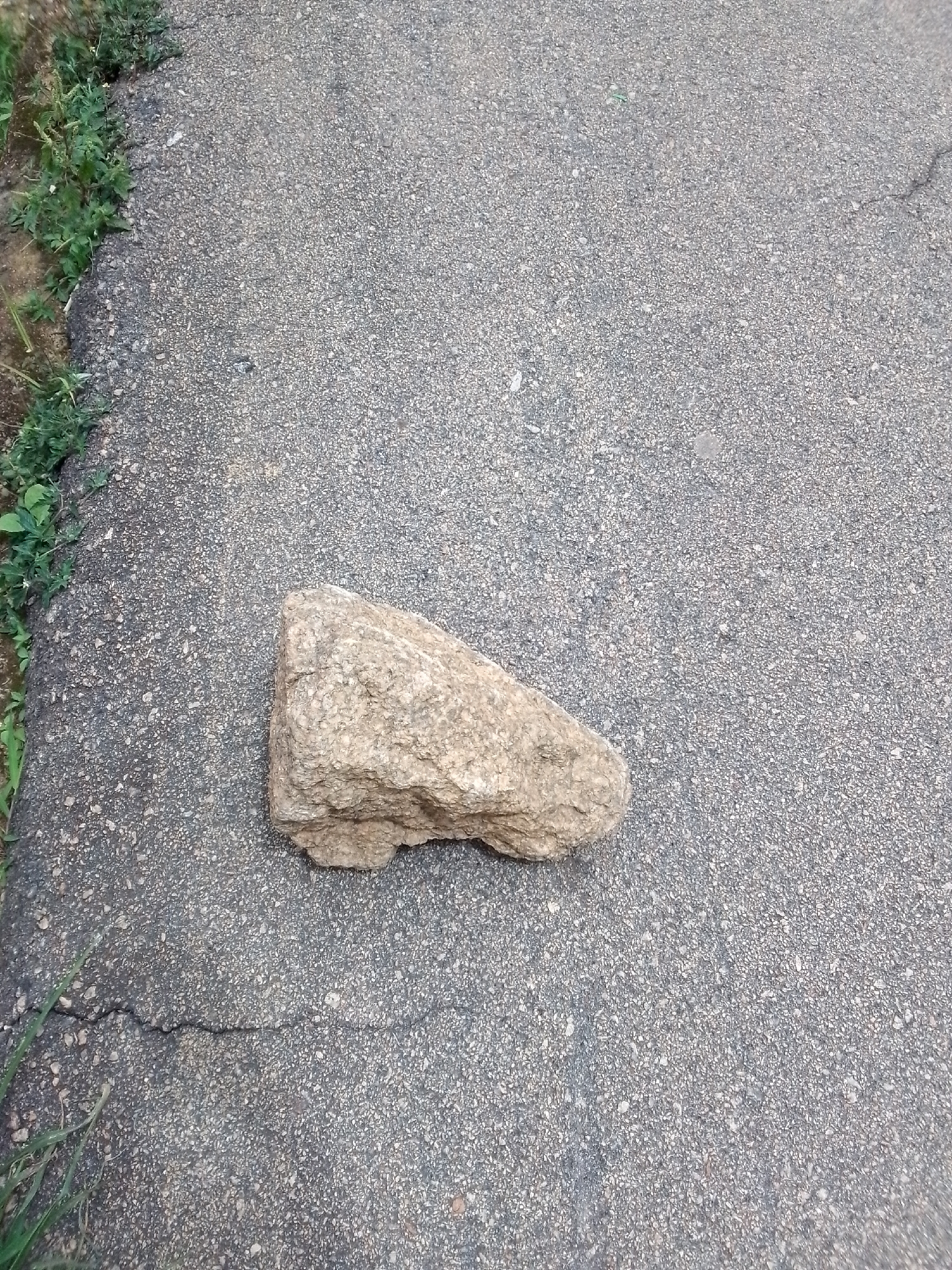 A single, light brown, rough-textured rock rests on a grey asphalt surface. The asphalt exhibits a fine aggregate texture and a visible crack runs diagonally from the bottom left to the middle of the image. To the left of the asphalt, a patch of green vegetation is present, with some sandy soil visible beneath it. The scene appears to be outdoors, possibly on a sidewalk or pathway in the city of Jos, Nigeria. No people or further objects are visible in the frame.
