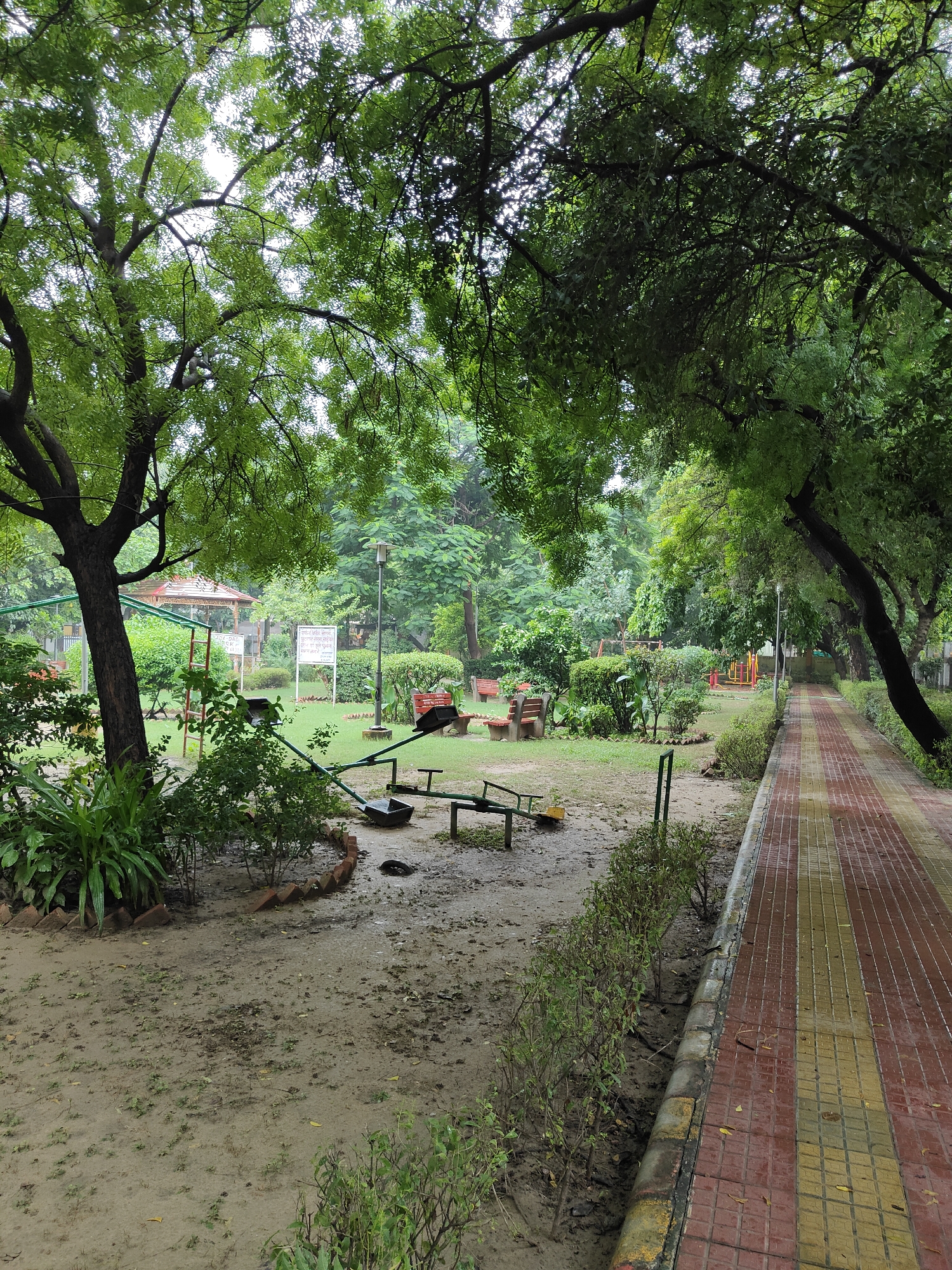 This image depicts a view of a park in Gurgaon, India, likely during the monsoon season given the wet ground and overcast sky. The foreground features a sandy, muddy patch of ground with scattered leaves and small green shoots. A section of a seesaw, made of green metal, is visible, with one of its seats resting on the ground. To the right, a paved walkway with red and yellow rectangular tiles runs along the edge of the park, bordered by a low hedge.
The middle ground of the park is characterized by a grassy area with several benches made of concrete and wood. A lamp post stands near the center of the park. Further back, there is a playground area with some colorful play structures, including what appears to be a small slide or climbing frame. A small pavilion or gazebo with a red-tiled roof is also visible in the background.
The park is densely populated with trees, whose branches and green foliage create a canopy overhead, filtering the light. The leaves of the trees are lush and green, suggesting a healthy and well-maintained environment. The overall atmosphere of the park is tranquil and somewhat subdued, perhaps due to the weather. There are no people visible in the image, making it appear deserted.