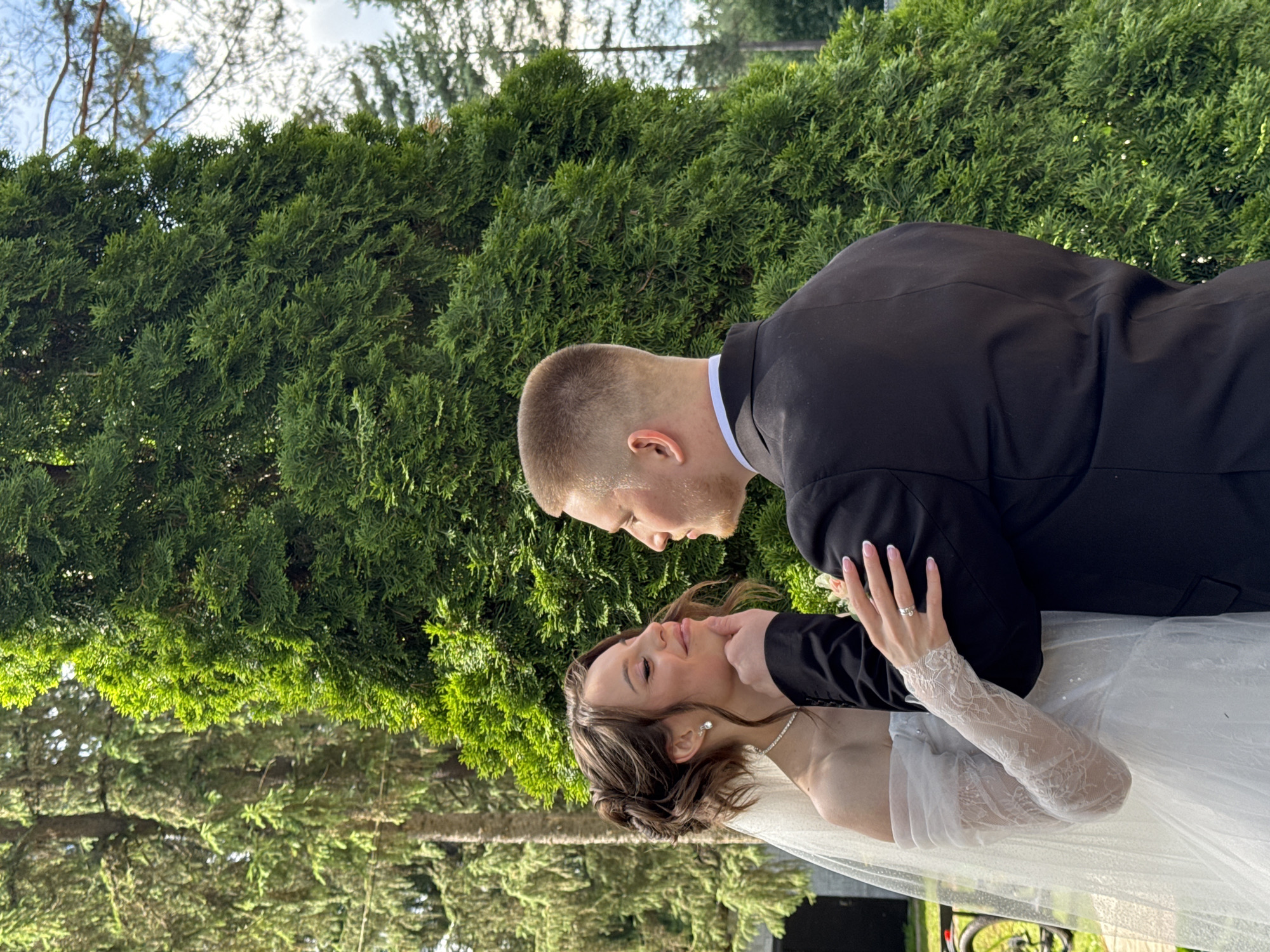 A young Caucasian couple, presumably newlyweds, are embracing outdoors in front of a large, lush evergreen tree. The woman, on the left, is wearing a white wedding dress with sheer lace sleeves and a veil. She has her head tilted up and her eyes closed, with the man's hand gently touching her chin. She is wearing a diamond necklace and stud earrings. The man, on the right, is wearing a black suit and white shirt. He is looking at the woman. The background features more trees and a cloudy sky, suggesting a natural or park-like setting. The image appears to be taken during daylight. There is no specific indication within the image to suggest the location is the city of Votkinsk, Russia.