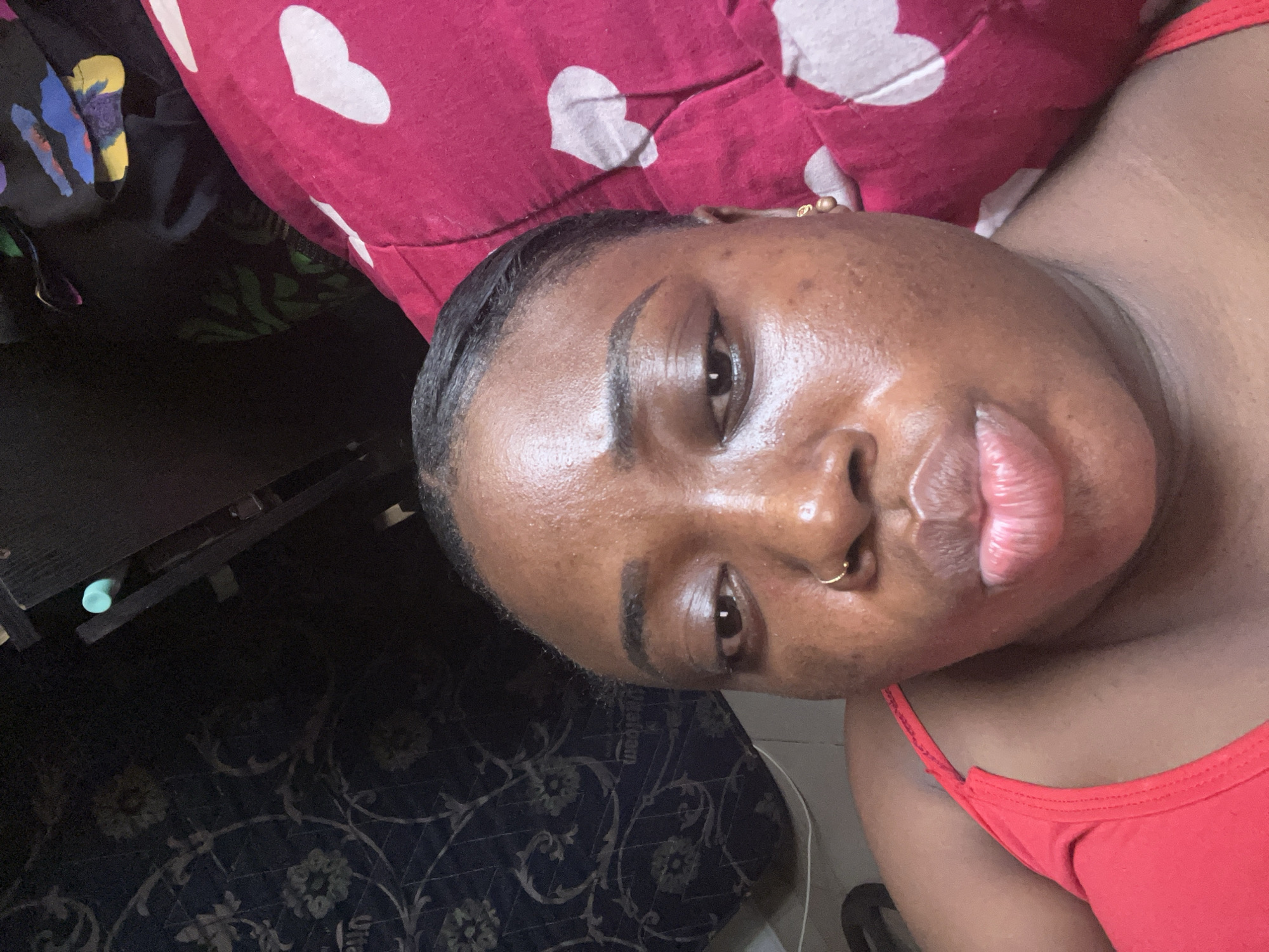 A medium shot shows a woman lying on a pillow with a red and white heart pattern. She has dark skin, dark hair styled back, and wears a gold earring and nose ring. Her expression is neutral. She wears a red tank top. Part of a desk and patterned rug are visible in the background. It is not possible to determine the location of Abuja, Nigeria, in the photograph.