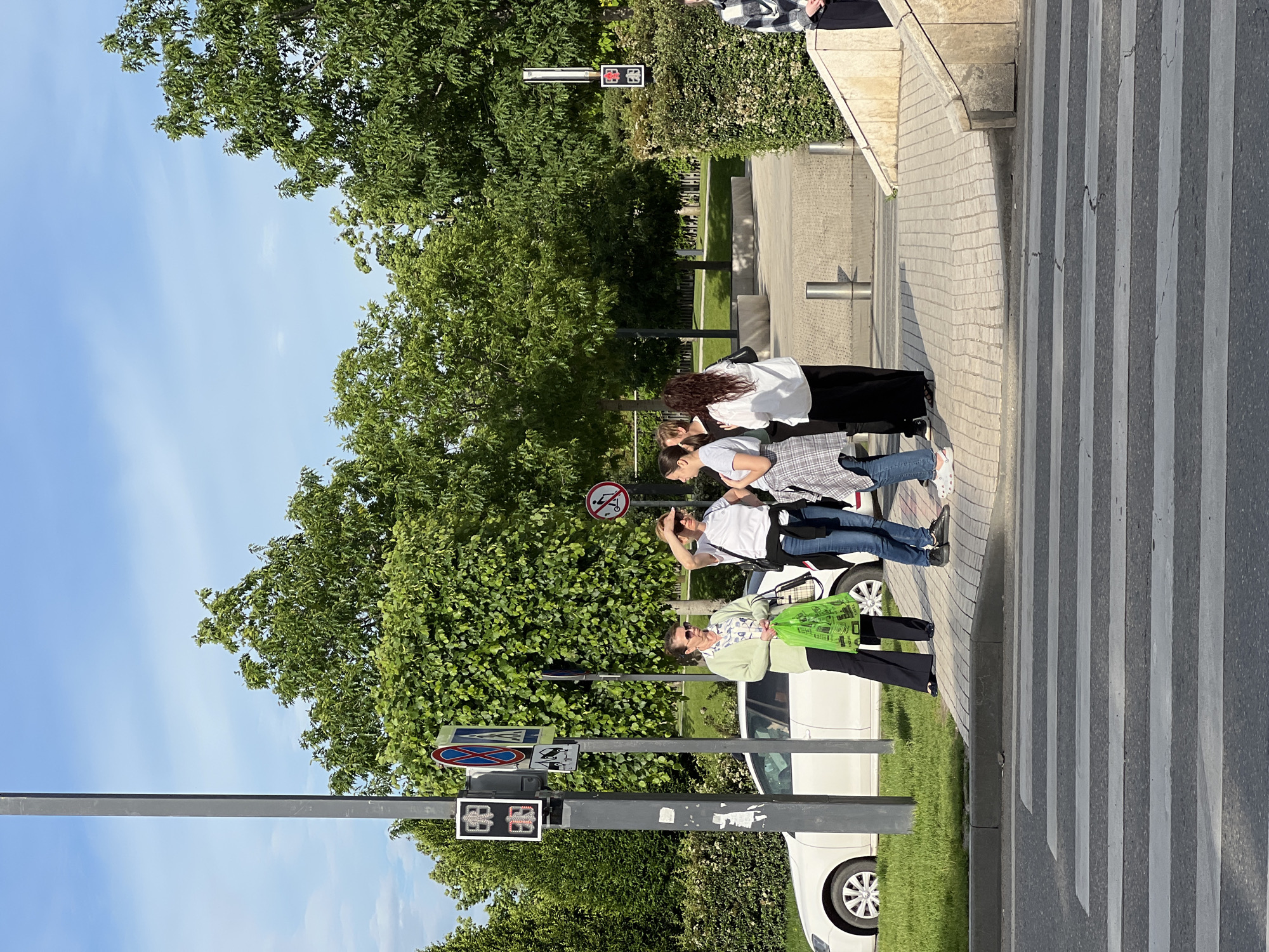The image shows a group of four people standing on a crosswalk in Krasnodar, Russia. They are positioned near a street sign and a traffic light. A white car is parked on the side of the street, adjacent to a grassy area. Trees and vegetation are visible in the background, partially obscuring the sky. The crosswalk has white stripes painted on the dark asphalt road. The pedestrian light shows red signal in both directions. The four people are waiting by the crosswalk. From front to back: An older lady with a light green jacket and a dark green shopping bag, a middle aged lady with a white shirt and blue jeans, and two ladies in white shirts and black pants.