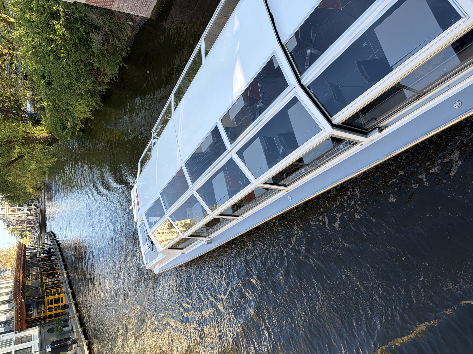 A white, multi-windowed canal boat, typical of those found in Amsterdam, navigates a tranquil canal. The boat is depicted from an elevated perspective, appearing slightly angled across the frame, with its bow pointing towards the upper left. Numerous large, clear glass windows line its cabin, suggesting it is a tour boat designed for passengers to observe the surroundings. Some windows appear partially open.
The water in the canal is dark but reflects sunlight, showing a rippled texture and a visible wake behind the moving vessel, indicating active transit. On the left bank of the canal, dense green foliage and trees line the water's edge, backed by a brick structure, possibly part of a bridge or embankment. On the right bank, further in the background, a series of buildings can be seen, one featuring prominent yellow accents, possibly a restaurant or cafe with outdoor seating along the water.
The scene is brightly lit by natural sunlight, casting reflections on the boat's white surfaces and the water, suggesting it is daytime, likely late morning or afternoon. No people are clearly visible on the boat or along the canal banks, and no legible text can be identified in the image. The overall impression is one of a peaceful, sunny day on the canals of Amsterdam.