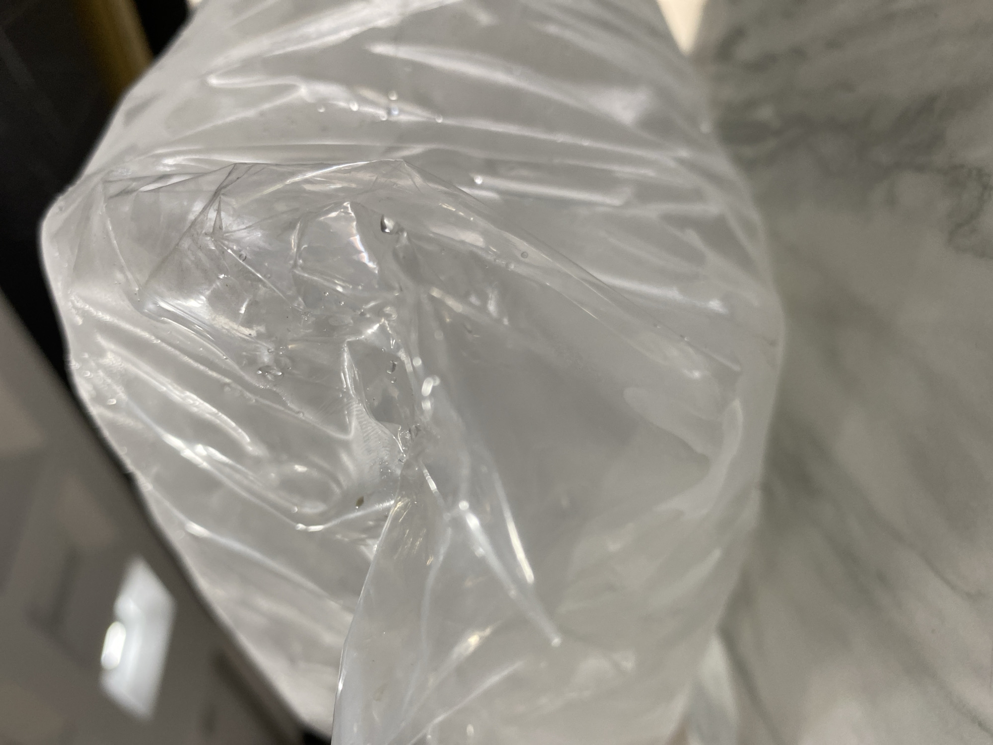 The image shows a close-up, low-angle shot of a translucent plastic bag filled with a white, opaque substance. The bag is wrinkled and appears to contain some moisture droplets on its surface. The lighting suggests an indoor environment with overhead fluorescent lights visible in the blurred background, which is characteristic of many commercial or public spaces in Banda Aceh, Indonesia. The focus is sharp on the texture and folds of the plastic bag, with the contents and the surrounding environment remaining indistinct. There are no people or discernible actions depicted.