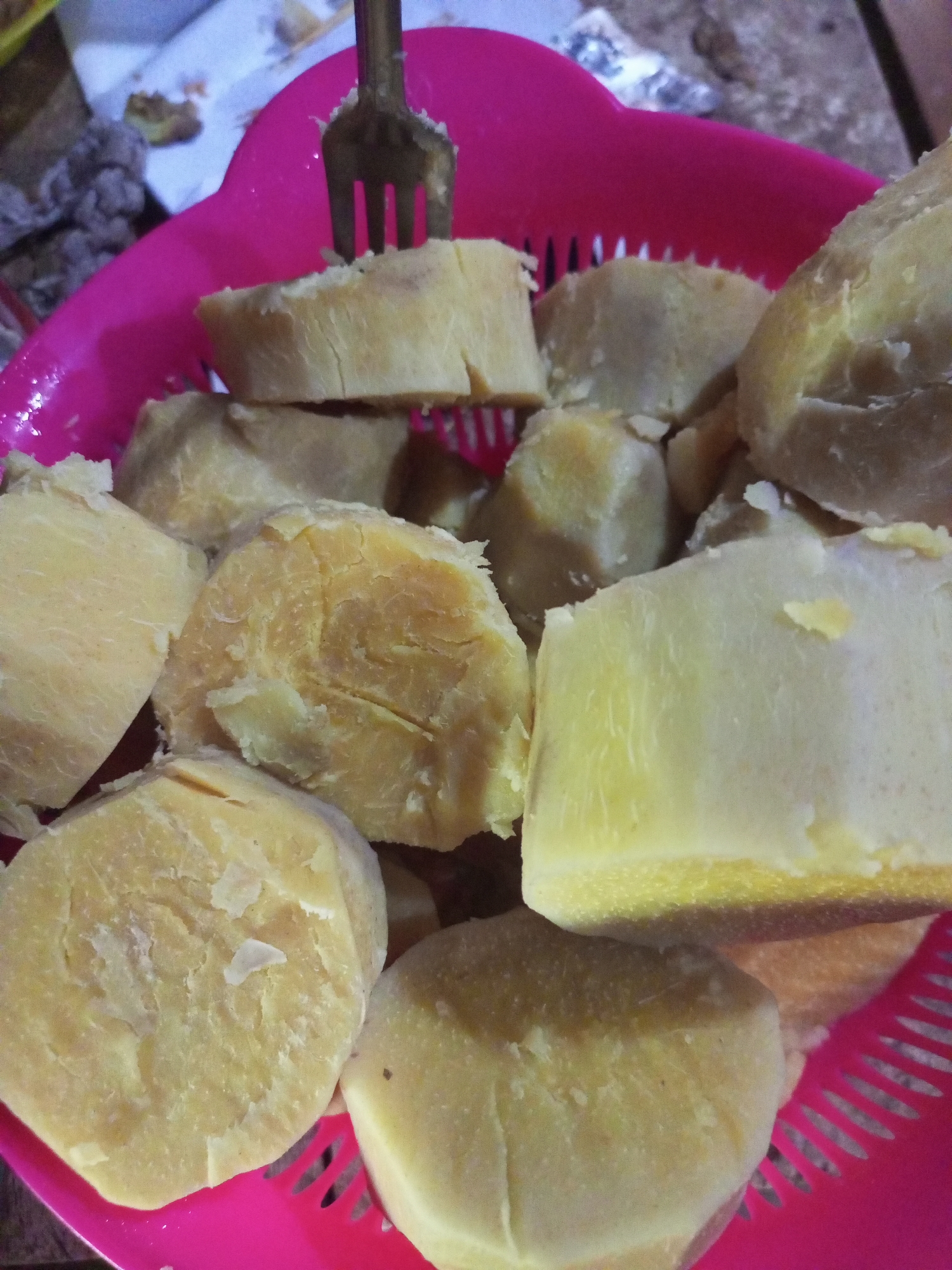 In a pink colander, cut pieces of boiled sweet potato are piled. A fork is visible, piercing one of the pieces of sweet potato. The sweet potatoes are a light yellow color and appear to be soft. The background of the image is blurry and suggests an outdoor setting, possibly a market stall, given the presence of other food items and woven materials. The lighting is natural and somewhat dim, indicating it may be overcast or late in the day.