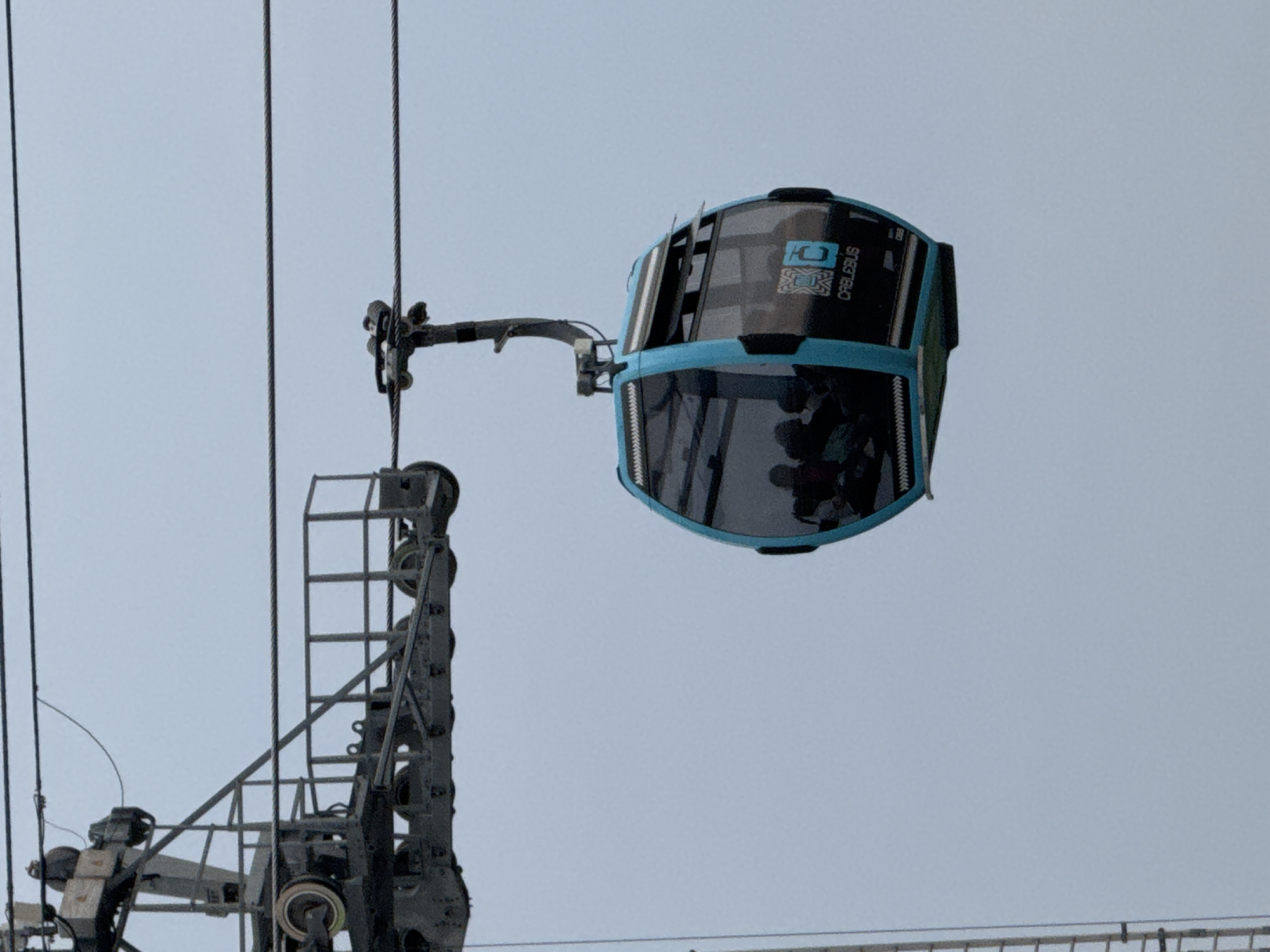 The image shows a Cablebus cable car in Mexico City, Mexico. The cable car is blue and black, and it is suspended from a cable. There are people inside the cable car. A steel structure with wheels supports the cable. The sky is overcast.