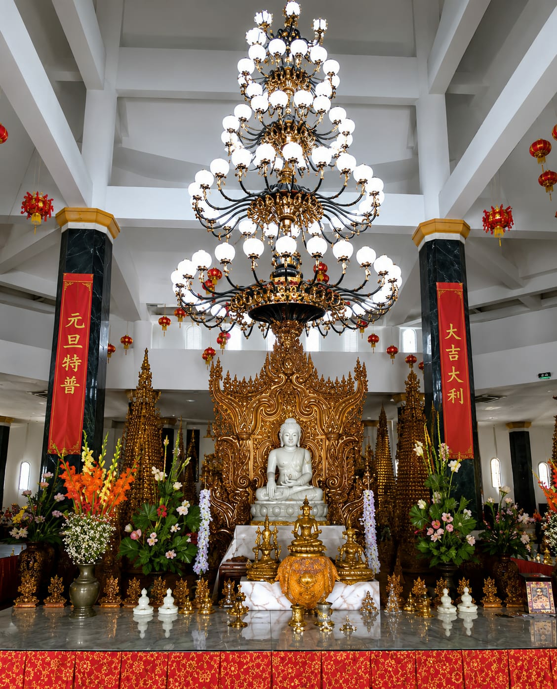 The image shows the interior of a Buddhist temple, likely located in Dolat Rayat, Indonesia, given the context. The central focus is a white Buddha statue seated on a golden, ornate throne. Flanking the Buddha are numerous golden statues of smaller Buddhas and decorative golden spires. In front of the throne, there are several white stupas and a large, golden, urn-like object. The altar is adorned with floral arrangements, including bright orange and pink flowers, and white flowers in vases. Two tall red banners with Chinese calligraphy hang from dark, marbled pillars on either side of the Buddha. The ceiling features a very large, multi-tiered, ornate chandelier with numerous lights. Red Chinese lanterns are visible hanging from the ceiling and the upper portions of the walls, suggesting a festive or celebratory occasion. The floor appears to be polished marble, reflecting the surrounding objects. The overall scene depicts a richly decorated and reverent space within a Buddhist temple.