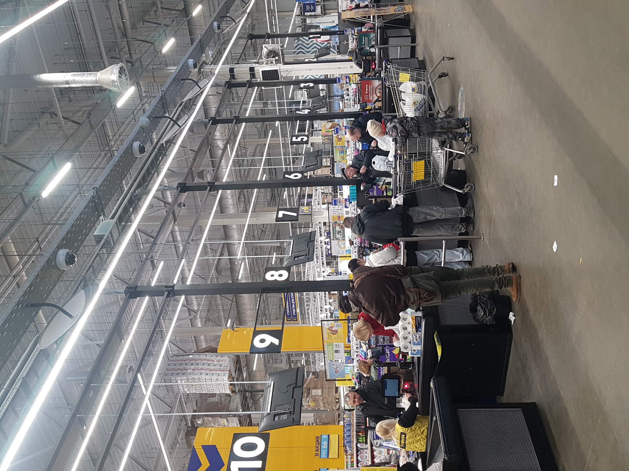 Here is a detailed description of the image:
The image shows a busy checkout area in a large supermarket, likely located in Saint Petersburg, Russia, based on the Cyrillic text and the "Lenta" branding visible. Multiple checkout lanes, numbered 3 through 10, are open with customers waiting in line. Most lanes have at least one shopper actively checking out or waiting their turn. Shopping carts filled with groceries can be seen in several queues.
The individuals are a mix of adults, possibly including children, dressed in casual attire. The cashiers, wearing yellow vests, are actively scanning items and interacting with customers at lanes 9 and 10. Various products are visible on the checkout shelves.
The setting is a large, open store with high ceilings and exposed industrial infrastructure, including pipes, ventilation ducts, and lighting fixtures. The lanes are marked with prominent yellow signs, numbered clearly in black. The floor appears to be polished concrete.