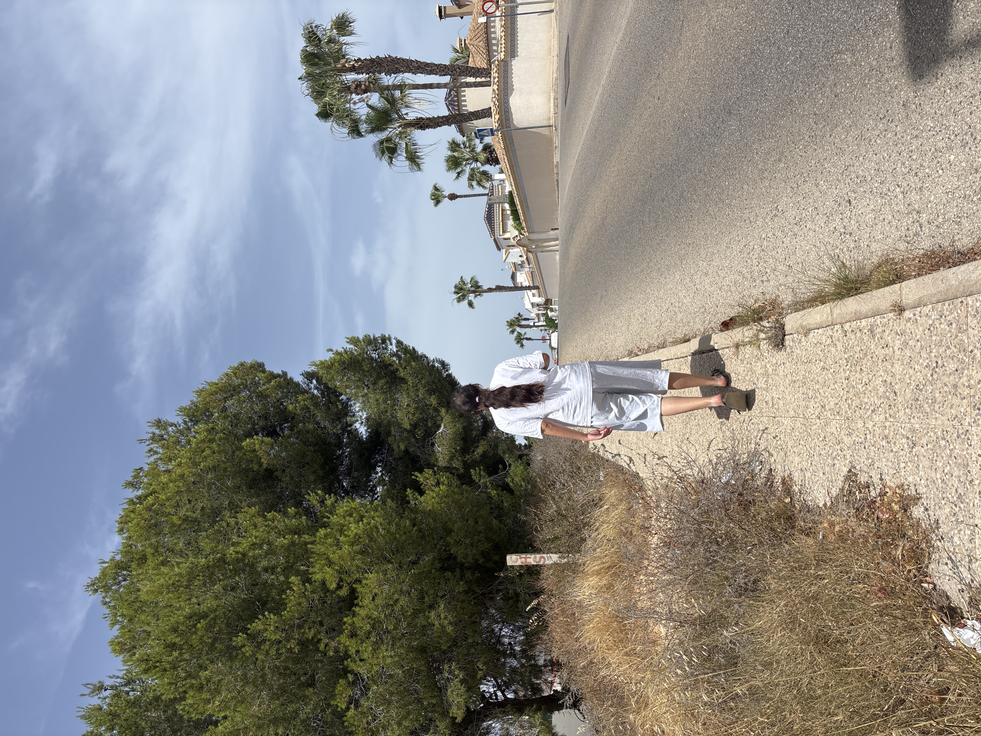 A person with long dark hair pulled back in a ponytail, wearing a white t-shirt and light-colored shorts, walks away from the camera on a paved sidewalk. The sidewalk runs alongside an asphalt road. To the left of the sidewalk is dry, overgrown vegetation and a large, mature evergreen tree. In the background, a row of white stucco houses with red tile roofs and palm trees line the opposite side of the road. The sky is blue with scattered white clouds. The scene appears to be in a residential area of Orihuela, Spain.