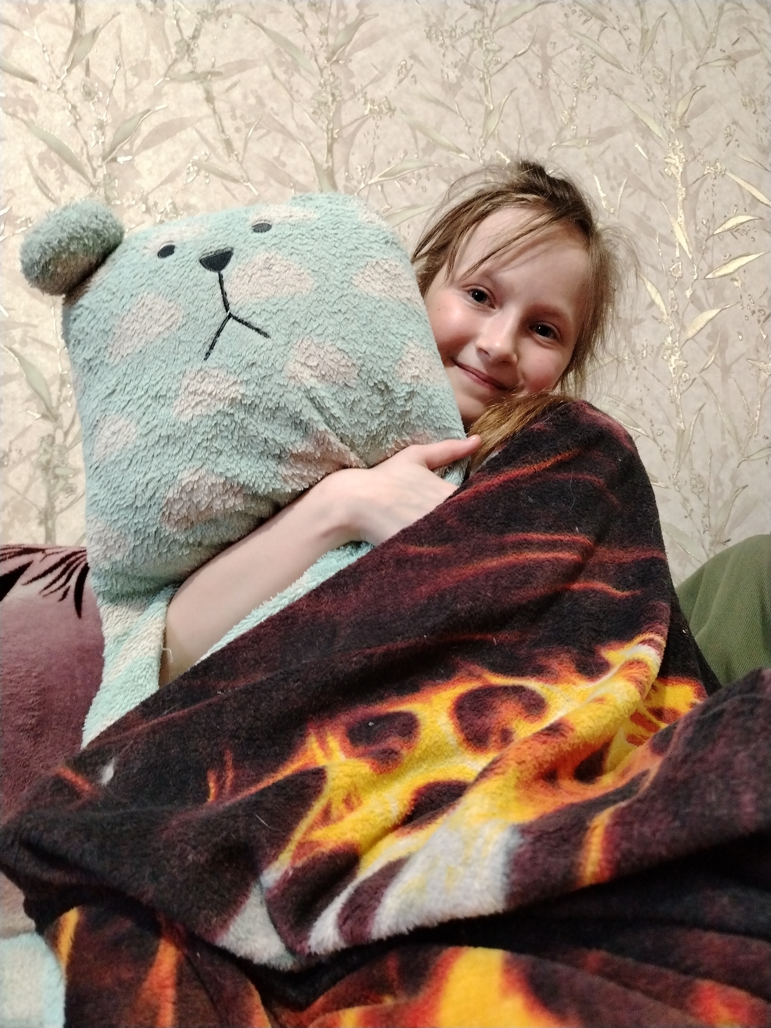 An indoor scene in Nizhnyaya Gar', Russia, shows a young female with light hair and fair skin, smiling directly towards the camera. She is partially covered by a dark-colored fleece blanket featuring a prominent pattern of red, orange, and yellow resembling flames. Her left arm is visible, embracing a light blue, plush, pillow-like object with two small black stitched circles for eyes and an inverted 'T' shape for a nose and mouth. A dark red cushion or armrest is partially visible on the far left. The background consists of a wall covered in light-colored wallpaper featuring a repeating pattern of reflective, golden-hued botanical elements. A small portion of olive-green fabric is visible on the far right.