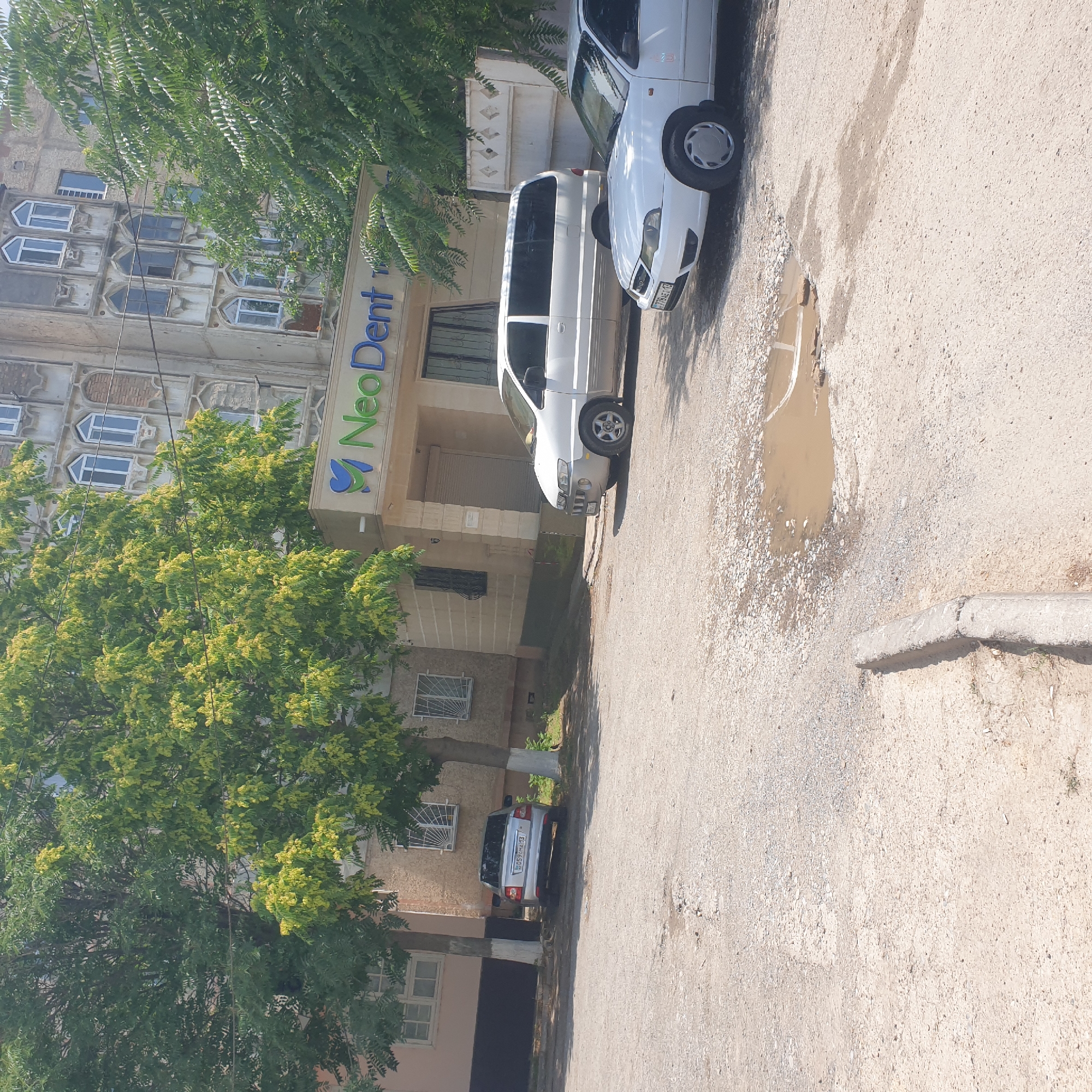 The image shows a street scene in Shymkent, Kazakhstan, with a building featuring the "NeoDent" logo prominently displayed. Several cars are parked on a rough, uneven asphalt surface with puddles and debris. Trees with green and yellow foliage are scattered throughout the scene, casting shadows on the ground. The building's architecture suggests it's an older structure, with arched windows and ornate details. The overall impression is of a typical urban environment with signs of wear and tear.