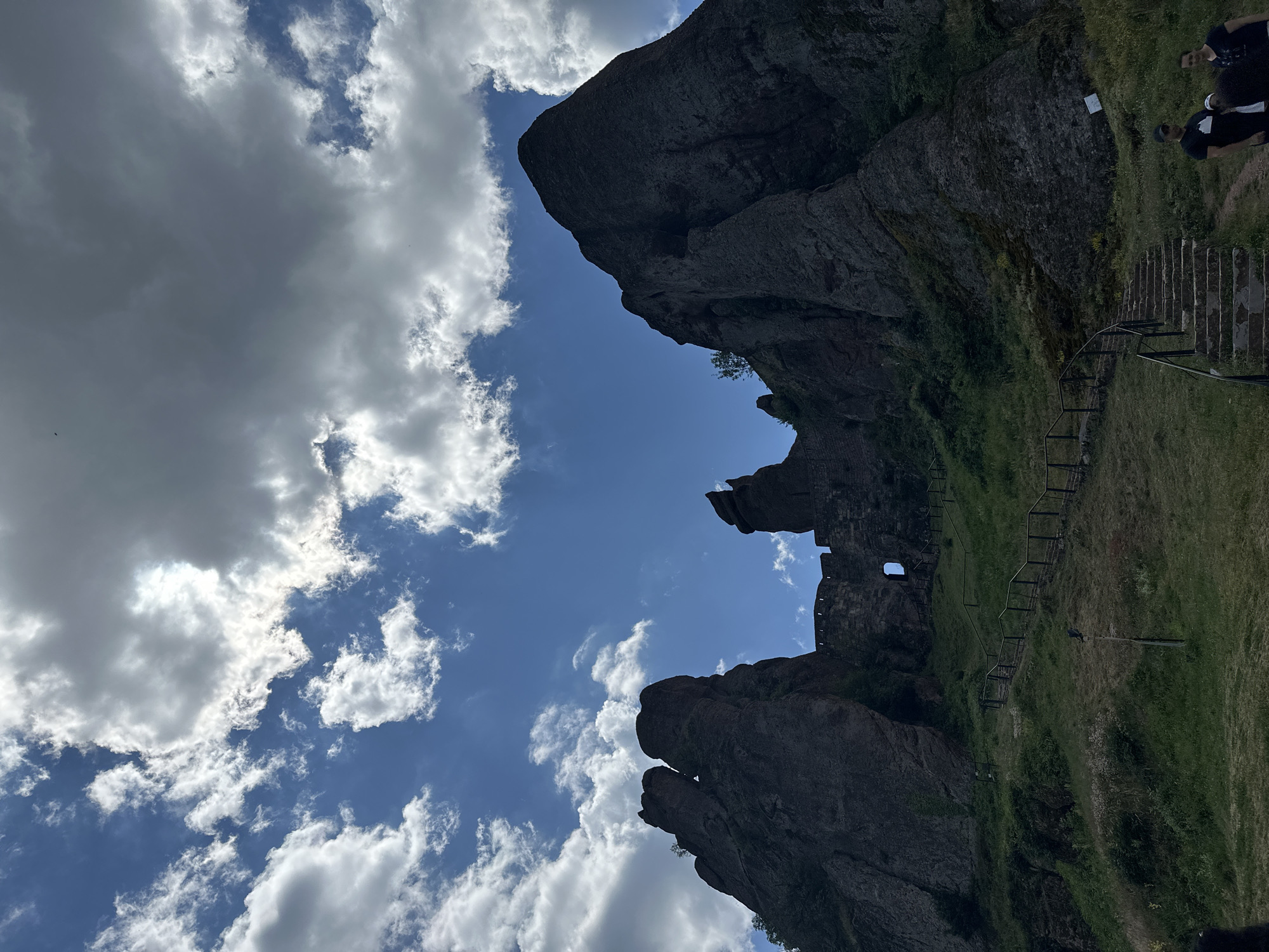 The image depicts a view of the Belogradchik Rocks, a UNESCO World Heritage Tentative List site in Belogradchik, Bulgaria. The foreground features a grassy slope with a stone staircase ascending to the right, bordered by a wooden railing. A second, less distinct wooden railing follows a path winding up the hill to the left. Several people are visible at the bottom right of the frame, appearing to be tourists.
Dominating the mid-ground and background are the massive, reddish-brown rock formations characteristic of the Belogradchik Rocks. These formations rise dramatically against a blue sky filled with white and gray clouds. Nestled among the rocks in the mid-ground, part of an ancient fortress is visible, constructed of stone blocks and featuring a small arched window. The fortress walls appear to be integrated with the natural rock formations. The overall scene conveys a sense of historical significance and natural grandeur.