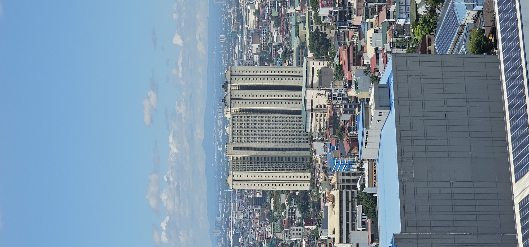The image depicts a panoramic view of the city of Makati, Philippines, under a clear blue sky with scattered white clouds. The scene is dominated by a dense urban landscape filled with numerous buildings of varying heights and architectural styles. In the mid-ground, a prominent, tall, light-colored residential building with numerous balconies stands out against the backdrop of the city. Surrounding this main structure are a multitude of smaller residential and commercial buildings, characterized by their diverse roof colors, including red, blue, and gray. Some buildings feature visible air conditioning units and antennas, indicative of a tropical urban environment.
In the foreground, a large, modern building with a gray facade and a light blue roof is partially visible. The rooftop of this foreground building shows a section with an array of solar panels, suggesting an emphasis on renewable energy. In the distance, a hazy mountain range is visible, providing a natural boundary to the cityscape. The overall impression is one of a bustling, densely populated metropolis with a mix of older and newer constructions. There are no people or vehicles clearly discernible in the image.