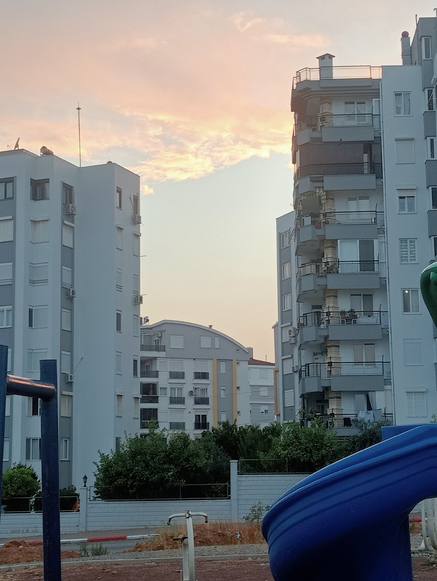 This is an eye-level shot of a residential area during sunset in Unknown, Unknown. The foreground is occupied by a blue plastic slide, a part of a playground. To the left, a blue metal pole stands vertically. Beyond the playground, there is a white fence with a red and white striped curb in front of it. Behind the fence, lush green trees and bushes are visible, with an old-fashioned street lamp peeking out from behind the foliage. Several apartment buildings dominate the background. The building on the left is tall and light blue, with many windows and air conditioning units. The building on the right is taller and predominantly white, with a more complex architectural design featuring multiple balconies and a grey facade. In the center, another apartment building, shorter and with a curved roof, is visible. The sky is filled with soft, pinkish-orange clouds, characteristic of a sunset. The overall scene depicts a quiet, residential environment at dusk.