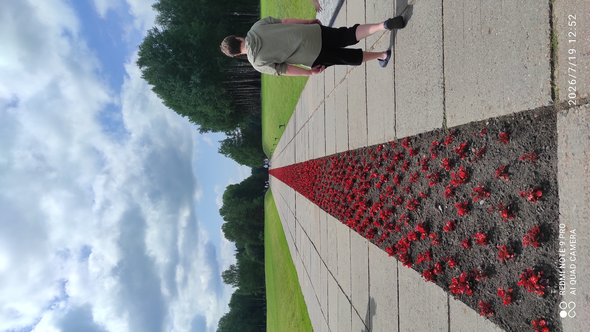 The image depicts a wide, paved pathway in an outdoor setting, lined with a long, narrow bed of vibrant red flowers. The pathway is constructed of large, rectangular concrete slabs. The flower bed runs down the center of the path, creating a striking visual contrast with the gray concrete. Beyond the pathway, to the left, is a neatly mown green lawn. To the right and in the distance, a dense line of tall, dark green trees forms a backdrop. The sky above is partly cloudy, with large, puffy white clouds against a clear blue sky. In the foreground, a person with short brown hair, wearing a light green hooded sweatshirt and black shorts, is walking away from the camera on the right side of the pathway. Further down the path, several other people are visible as small figures walking in the distance. The timestamp "2026/7/19 12:52" and the text "REDMI NOTE 9 PRO AI QUAD CAMERA" are present in the lower corners of the image, suggesting it was taken with a mobile phone. The scene appears to be a park or memorial site, possibly in Каменскі сельскі Савет, Belarus, given the context provided.