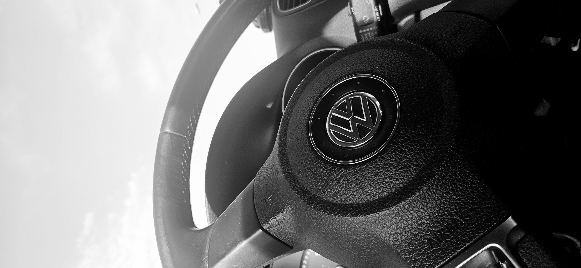 This black and white image captures a close-up, low-angle view of the interior of a car, specifically focusing on the steering wheel. The steering wheel, made of textured black material, occupies the central and right portions of the frame. In its center, a prominent circular emblem features the metallic silver Volkswagen (VW) logo against a dark background, surrounded by small reflective details. Below the logo, faint embossed text reads "AIRBAG".
To the upper right, a control stalk extends from the dashboard area, displaying various settings and text such as "ON/OFF", "RESET", "HIGH", "LOW", and "REAR", indicating functions like wipers or cruise control. A car air vent is partially visible in the upper left background. The left side of the image is dominated by a bright, almost overexposed white area, suggesting a daytime sky or bright outdoor conditions seen through the windshield. The high contrast lighting highlights the textures of the steering wheel and creates reflections on the metallic logo. The overall setting is inside a vehicle in Plovdiv, Bulgaria. No individuals are present, and no particular activity is depicted beyond a static view of the car's interior.