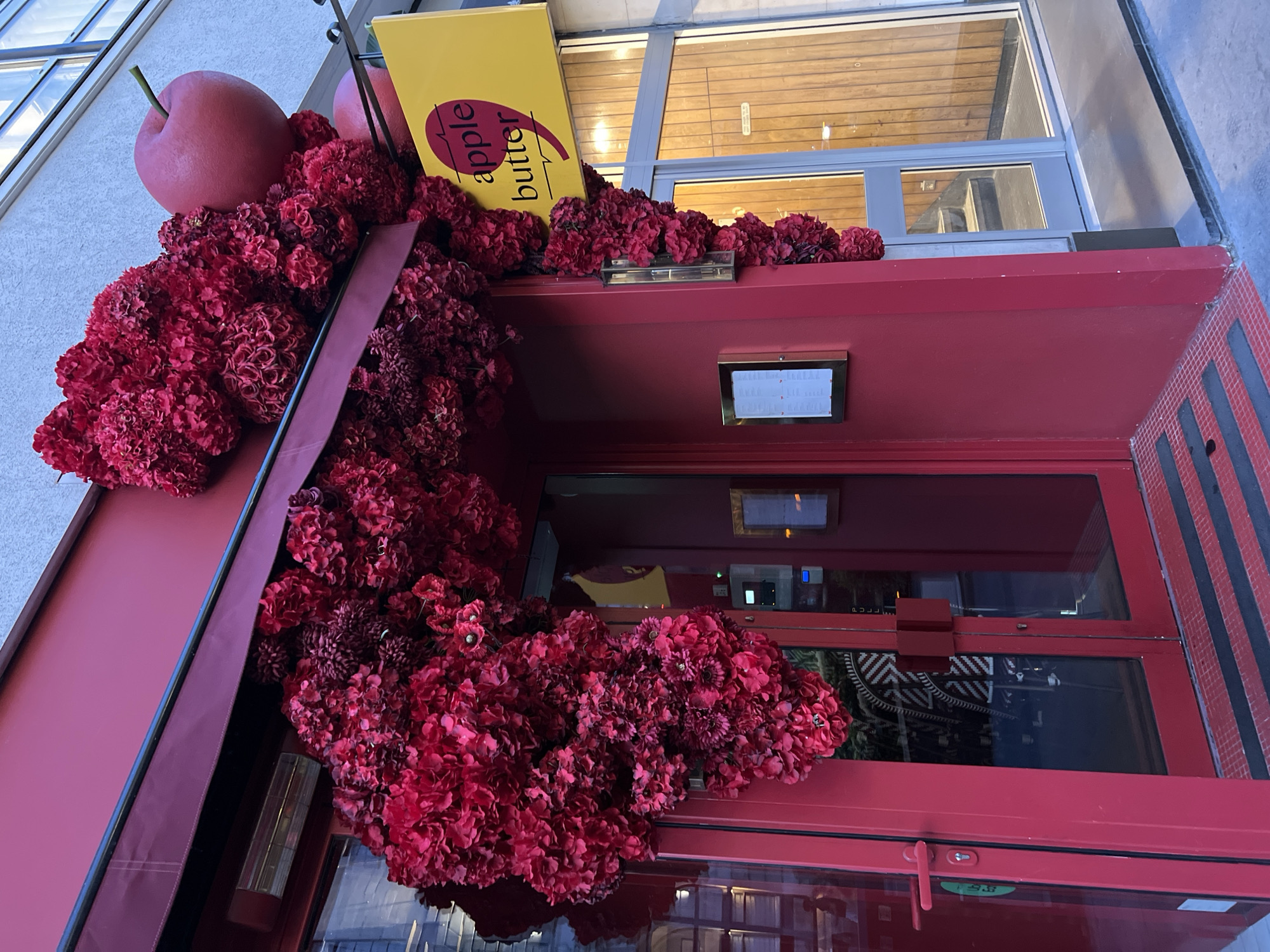 The image shows the exterior of a building, likely a shop or restaurant, decorated with an abundance of deep red floral arrangements. The entrance features double red doors with glass panels. Above the entrance, a red awning extends outwards, also adorned with the same red flowers. A yellow sign with the text "apple butter" in dark lettering is mounted above the entrance to the right. Two large, round, red decorative apples are placed on top of the floral arrangement, near the sign. The building's facade is painted red. To the right of the entrance, a glass door leads into the building, revealing a wooden-paneled interior with stairs visible. The ground in front of the entrance has a textured surface with red and black stripes. The overall impression is festive and eye-catching, with the red floral decorations creating a dramatic and vibrant display.