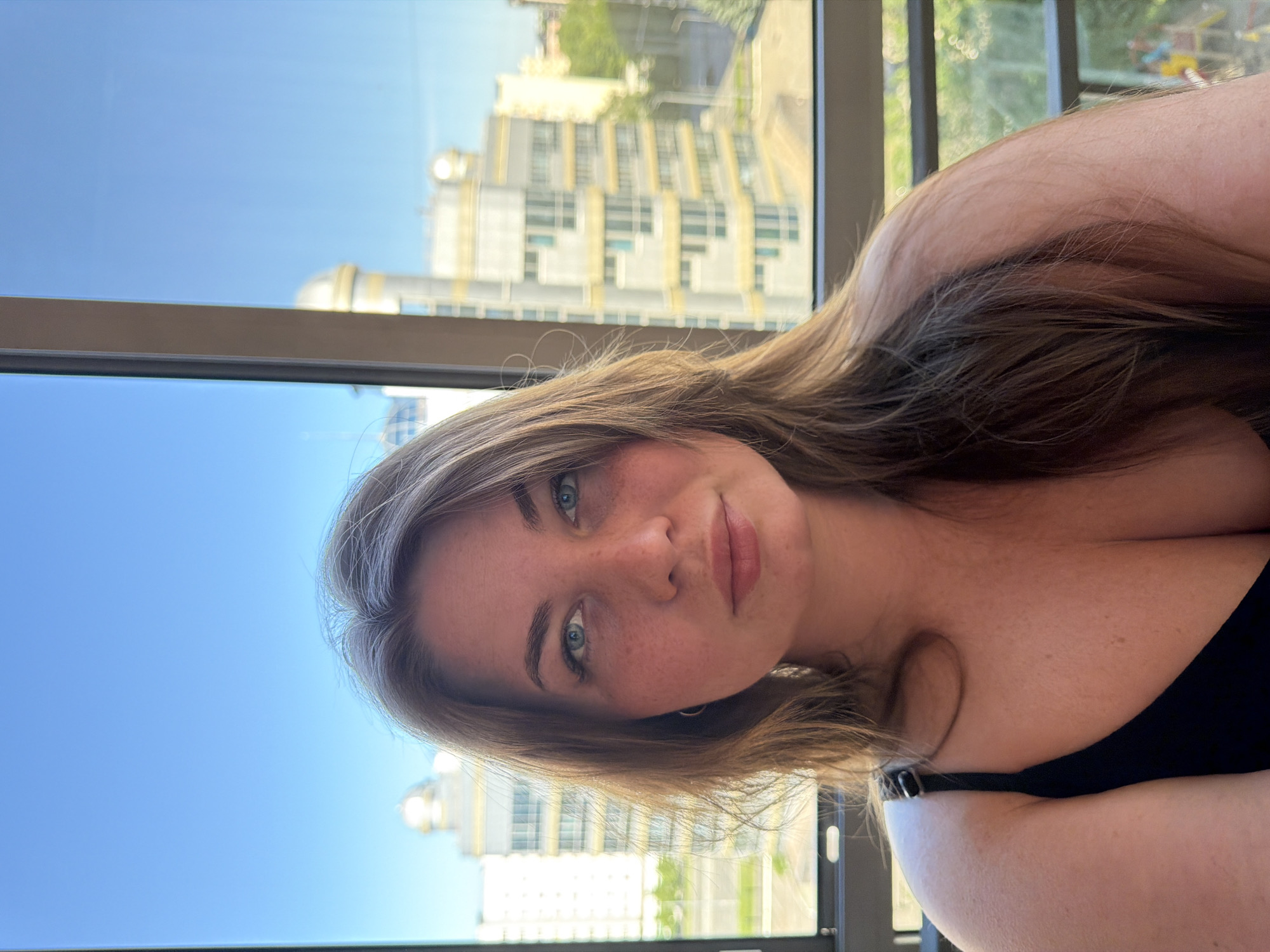 A young adult female with long brown hair, blue eyes, and freckles on her face looks directly at the camera. She is wearing a black top, with a strap visible on her left shoulder and cleavage exposed. The scene is set indoors, looking out through a large window, revealing a clear blue sky and city buildings under bright daylight. In the background, prominent multi-story buildings are visible. Specifically, a large architectural structure with distinct golden dome-like elements on its rooftop is clearly identifiable as a portion of the Kazan Family Center (also known as the Kazan Wedding Palace or 'The Bowl') in Kazan, Russia. Below the buildings, green foliage and what appears to be a section of a children's playground are visible.