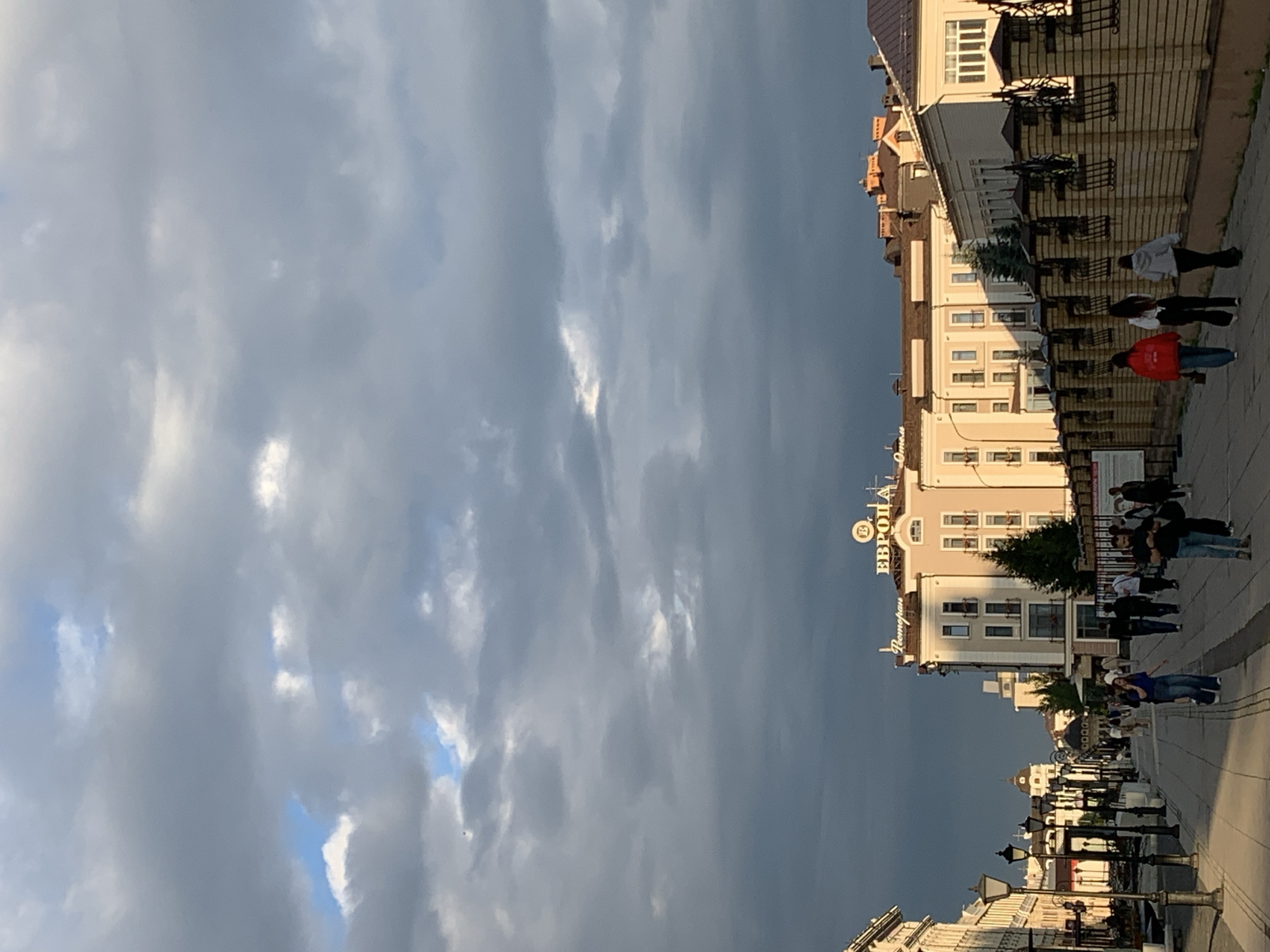 This is a vertical, eye-level, full-shot image of a street scene in Kazan, Russia, likely on Bauman Street, during the late afternoon or early evening. The sky is overcast with dramatic, dark clouds in the upper portion of the frame, while patches of blue sky and lighter clouds are visible towards the top. The sun, low in the sky, casts a warm, golden light on the buildings and the street from the right side, creating long shadows.
The street is paved and appears to be a pedestrian walkway lined with lampposts and shops. Several people are walking along the street, some individually and some in small groups. They are dressed in casual clothing. On the right side of the street, there is a large building with the Cyrillic sign "ЕВРОПА" (Europe) in illuminated letters above the entrance, suggesting it is a hotel or a commercial establishment. Adjacent to this building is a decorative brick wall with wrought-iron accents.
On the left side of the street, there are more buildings with architectural details typical of historic European cities. The perspective leads the viewer's eye down the street, towards a distant view of more buildings and a dome. The overall atmosphere is one of a bustling city street with transitioning weather, as suggested by the dark clouds and the warm sunlight.