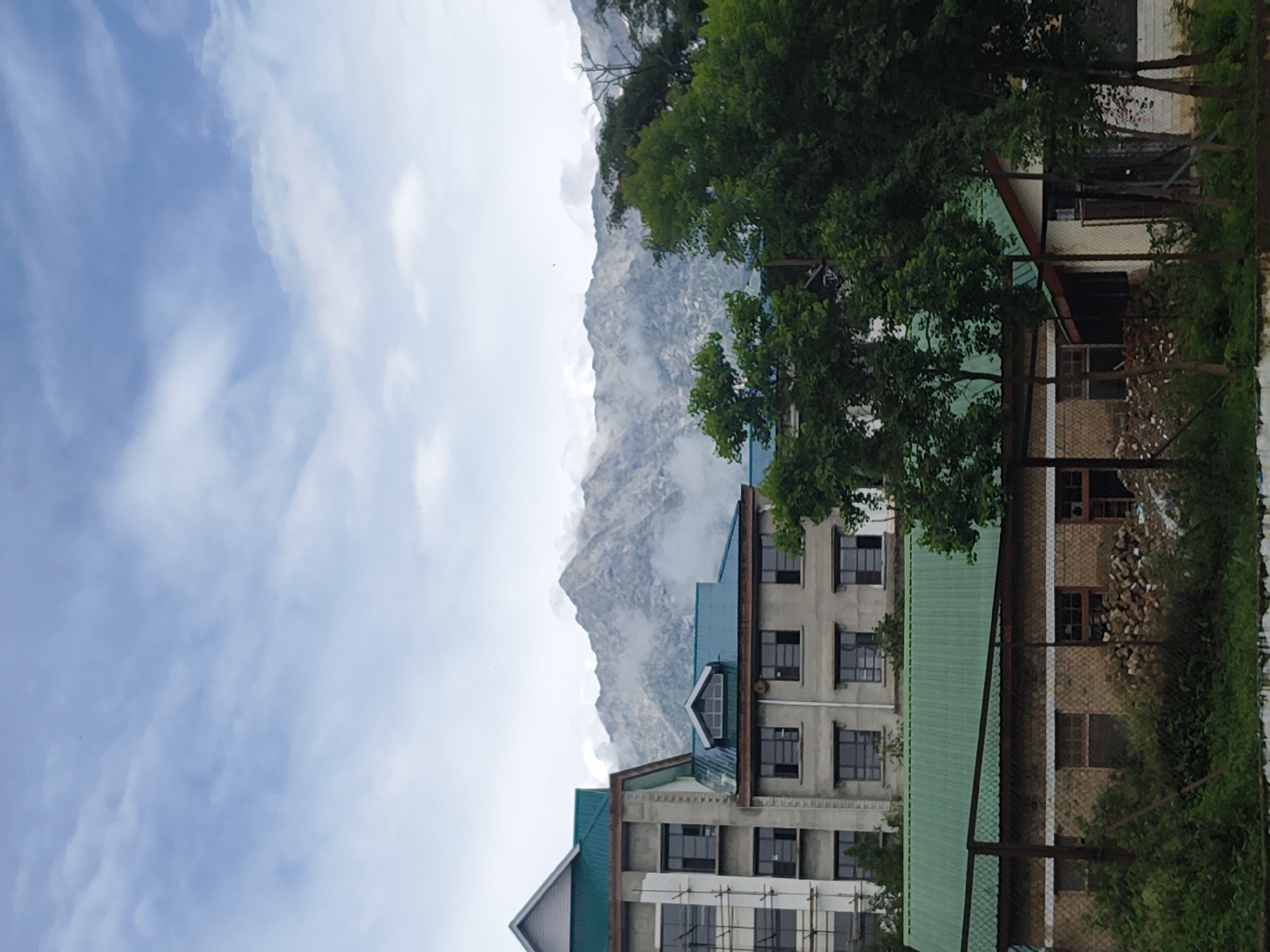 The image depicts a scene in Dharamshala, India, featuring a multi-story building with a teal-colored roof, constructed with concrete and glass windows, set against a backdrop of misty mountains. In the foreground, a single-story structure with a similar teal roof is visible, constructed of brick. The area is characterized by lush green trees and vegetation surrounding the buildings. The sky is overcast with clouds, indicating a possibly hazy or rainy day. No people are visible in the image. The scene appears to be part of an institutional or residential complex, given the architecture of the buildings. The presence of scaffolding on one of the buildings suggests ongoing construction or renovation work.