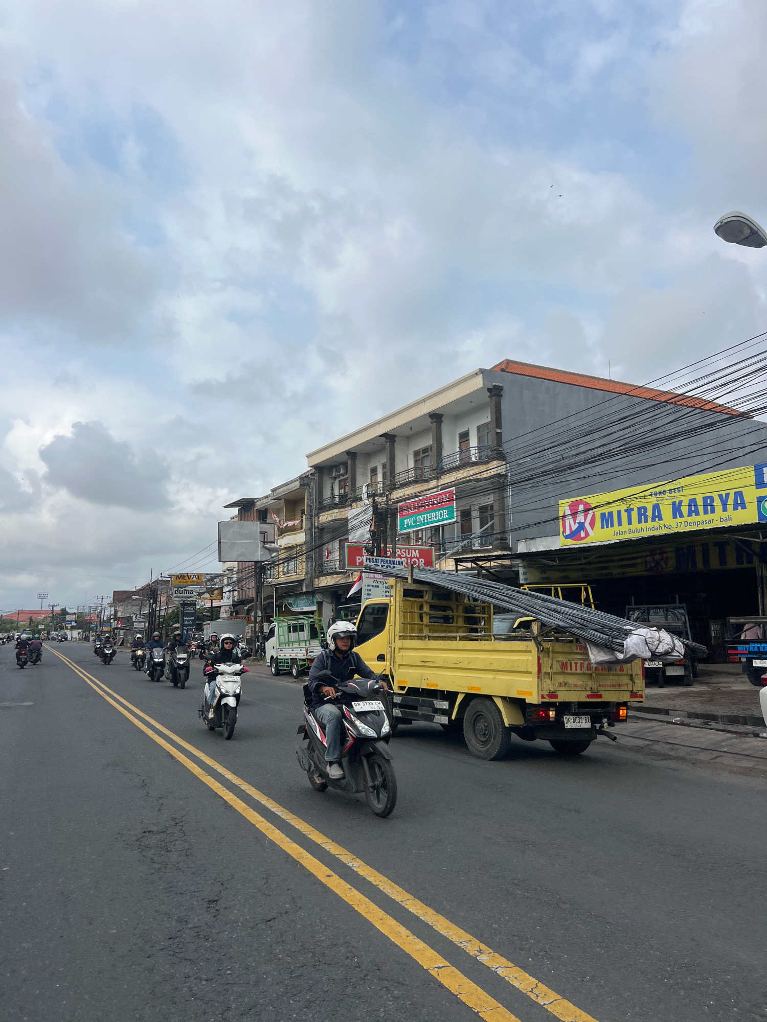 The image depicts a street scene in Denpasar, Indonesia, under an overcast sky. Multiple motorcycles are in motion on a paved road with double yellow lines. On the right side of the road, several commercial buildings are visible, featuring signs for businesses such as "MITRA KARYA" and "PVC INTERIOR". A yellow flatbed truck loaded with metal pipes is parked on the side of the road. People are riding the motorcycles, many wearing helmets. Electrical wires are strung across the scene above the buildings. The overall impression is a typical urban street environment during daylight hours.
