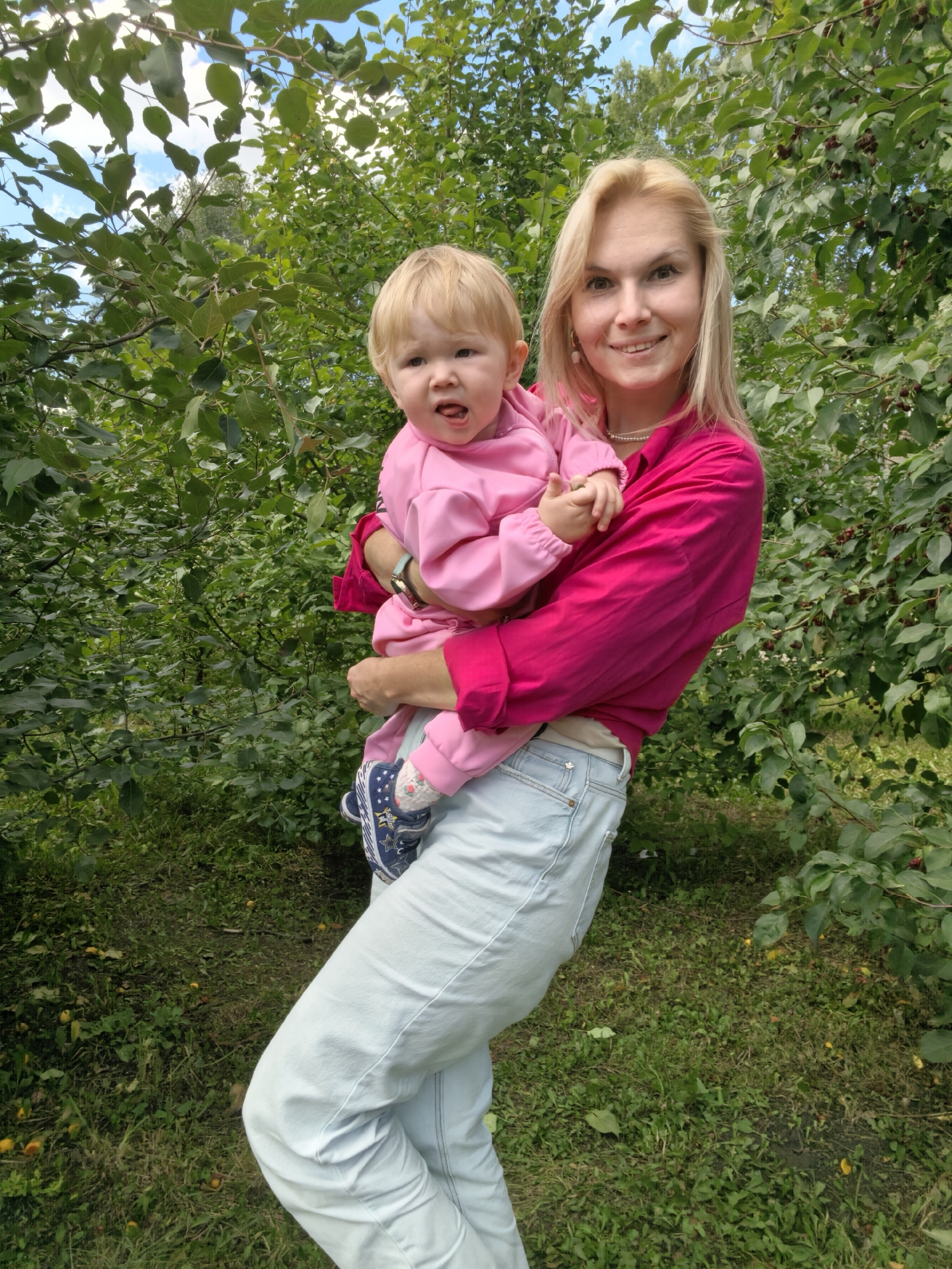 In Sterlitamak, Russia, a blonde woman in a bright pink shirt and light blue jeans holds a baby dressed in pink. The woman is smiling at the camera, while the baby looks slightly away with its mouth open. They are positioned in front of lush green foliage, with some fallen fruit visible on the grassy ground.