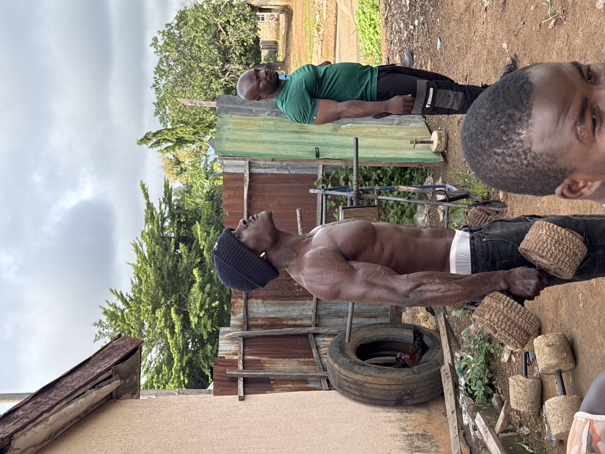 In Akwanga, Nigeria, a group of men are engaged in a workout session in what appears to be an outdoor gym set up in a yard. In the foreground, a muscular man wearing a black beanie and dark pants is performing bicep curls with two homemade dumbbells. He is shirtless, showcasing his physique. To his right, another man in a green t-shirt and black pants stands, observing. Behind them, a rusted corrugated metal fence serves as a backdrop, with a large truck tire leaning against a building on the left. The ground is dirt and debris, with more makeshift weights scattered around. Trees and vegetation are visible in the background, suggesting a rural or semi-urban setting. The sky is overcast, indicating a cloudy day.