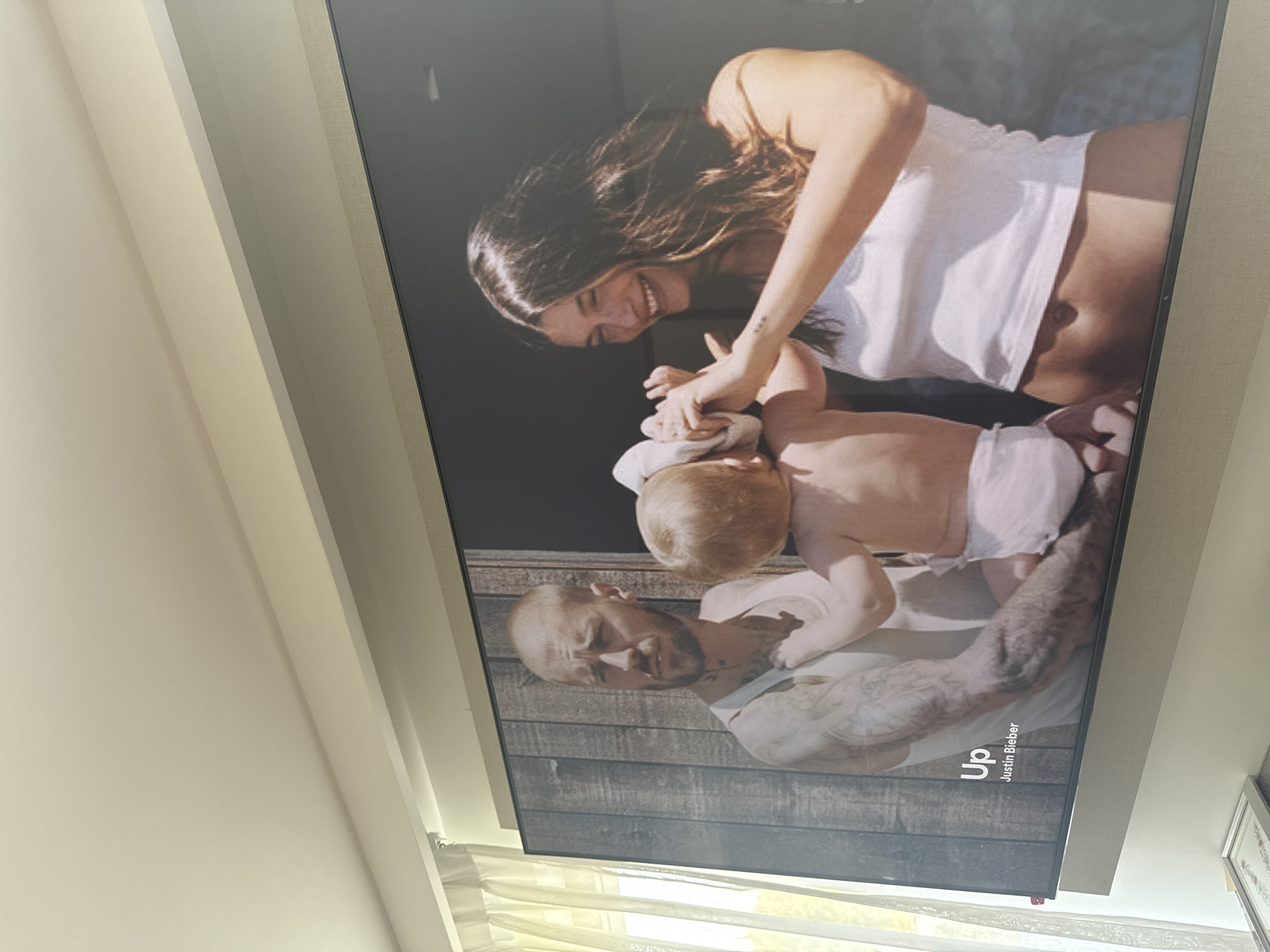 A large, vertically oriented television screen displays a tender family scene within a room located in San Bernardino Tlaxcalancingo, Mexico.
On the screen, a joyful woman with long, dark hair, wearing a white tank top, leans in from the upper right to playfully interact with a baby, holding its hands. Her expression is one of bright laughter. In the lower left of the screen, a man with a shaved head and extensive tattoos on his arms and neck holds the baby, looking directly towards the viewer with a gentle and pensive expression. The baby, with light-colored hair, is clad in a simple light-toned garment or diaper and is positioned centrally between the two adults. The background of the displayed image features rustic wooden paneling.
In the bottom right corner of the screen, the text "Up" and "Justin Bieber" is visible, indicating the content is likely a music video or promotional material.
The physical setting surrounding the screen includes a plain, light-colored ceiling and walls. Below the screen, bright, diffused daylight streams in through light-colored curtains or blinds, suggesting it is daytime. A portion of a framed picture is also visible on the right side of the room, adjacent to the screen. The scene conveys a heartwarming moment of familial connection presented on a modern display, situated within a simple and brightly lit indoor environment.