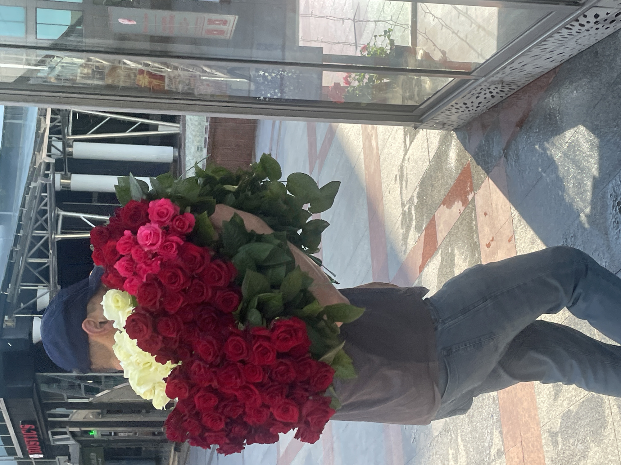 An individual, wearing a dark t-shirt and blue jeans, is holding a large bouquet of red and yellow roses. The person is also wearing a dark blue baseball cap. The scene appears to be outdoors in an urban environment. In the background, there is a building with the word "RUSTIC'S" visible on its facade, suggesting it could be a shop or restaurant. Adjacent to the person and bouquet is a large glass storefront, with some potted plants visible inside and reflecting the surroundings. The ground is paved with stone tiles, some of which appear wet. The lighting indicates it is daytime. The context of Sochi, Russia is not discernible from the visual information present in the image.