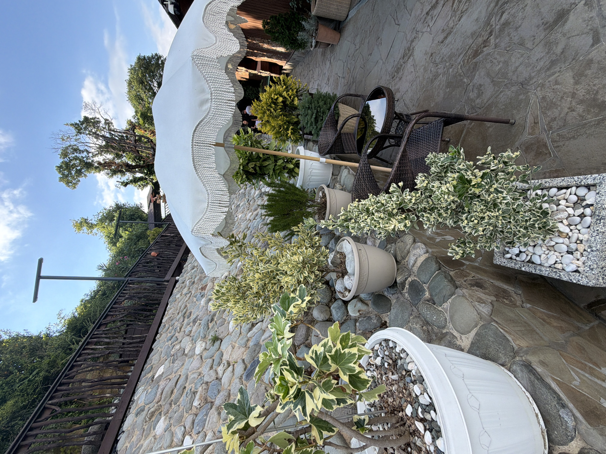 The image depicts an outdoor seating area on a stone-paved pathway, likely in a restaurant or cafe. A large white umbrella with a fringed edge provides shade over two brown wicker chairs and a small round table. Several potted plants, including a variegated ivy and a small bushy plant, are arranged around the seating area and on a stone retaining wall. The background features a wooden fence, lush greenery, and a blue sky with scattered clouds. In the distance, two individuals can be seen walking down the pathway. The scene suggests a relaxed and inviting atmosphere, possibly in the city of Nizhnyaya Shilovka, Russia, given the natural elements and outdoor setting commonly found in such locations.