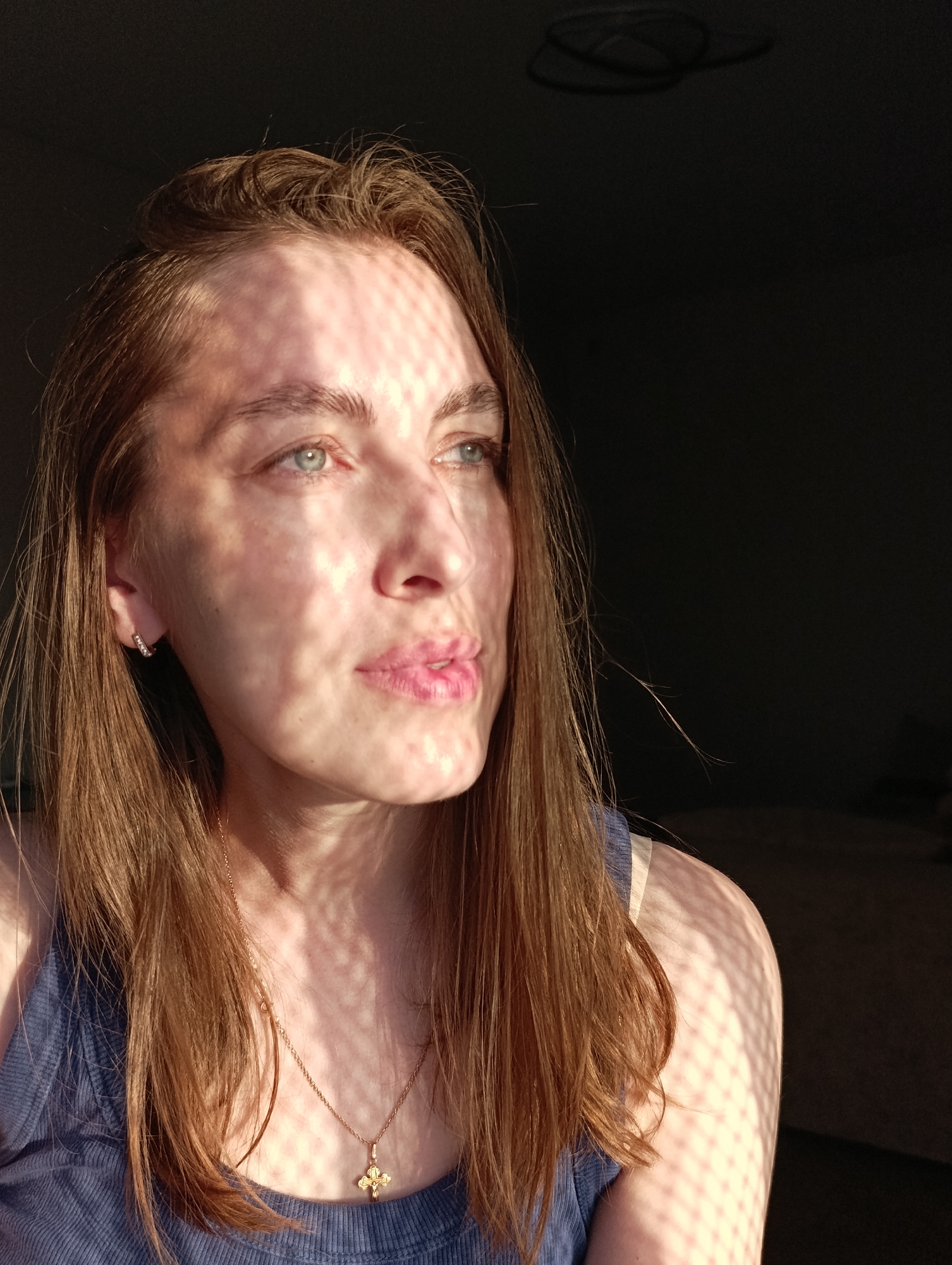 A young Caucasian woman with long, auburn hair and blue eyes is pictured in a close-up, head-and-shoulders shot. She is wearing a blue tank top and a delicate gold necklace with a cross pendant. The woman's face is partially illuminated by dappled sunlight, creating a criss-cross pattern of light and shadow across her features. She is looking off to the right, her lips slightly parted. The background is dark and blurred, suggesting an indoor setting. The presence of a ceiling fan is visible in the upper portion of the image. The scene is likely from a residential apartment in Chaikovsky, Russia, given the ambient lighting and common household objects.