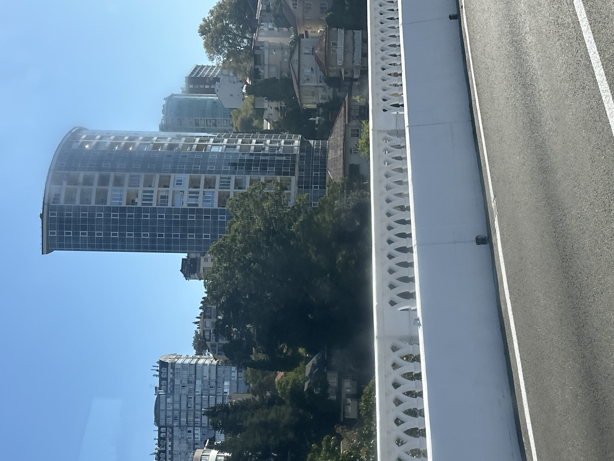 The image captures a cityscape with a clear blue sky. In the foreground, a white bridge railing with a decorative, arched pattern is visible above a white concrete barrier. A paved road with a white line runs alongside the barrier. Behind the bridge, several buildings of varying heights and architectural styles are present.
To the left, a large, multi-story building with a grey facade and numerous windows stands out. Adjacent to it, and slightly behind, are other buildings with a similar modern aesthetic. In the center of the image, a tall, curved skyscraper with a glass facade dominates the view. To the right of this skyscraper, other high-rise buildings are visible, some appearing to be under construction or with tinted glass.
Nestled among the modern buildings and rising hills are older residential houses with tiled roofs and balconies, suggesting a mix of development. Lush green trees and vegetation are scattered throughout the scene, particularly in the mid-ground, softening the urban landscape.
This scene is characteristic of the coastal areas of Sochi, Russia, specifically the area around the Khosta district, known for its modern high-rise hotels and residential complexes interspersed with older architecture and abundant greenery along the Black Sea coast. The elevated perspective suggests the photo was taken from a vehicle moving along a road or bridge.