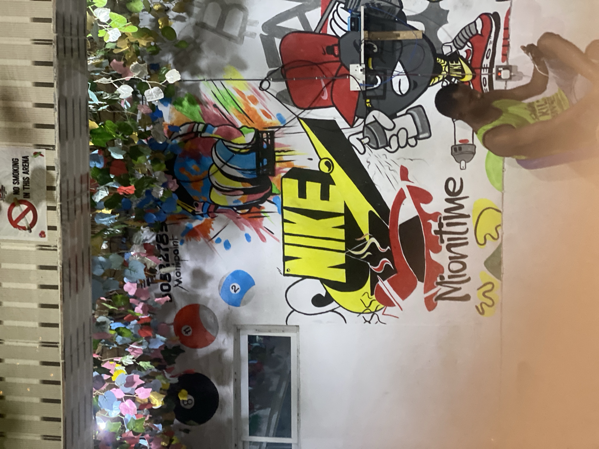 The image shows a mural on a wall in Badagry, Nigeria. The mural features cartoonish imagery including spray paint cans, a shoe, a lightning bolt, the word "Nike" stylized with a lightning bolt in place of the swoosh, and the words "Mionitime." There are also painted billiard balls. A window is visible below the mural. A person is sitting to the right of the mural. A "No Smoking in this area" sign is visible on the left side of the image, next to decorative plants.