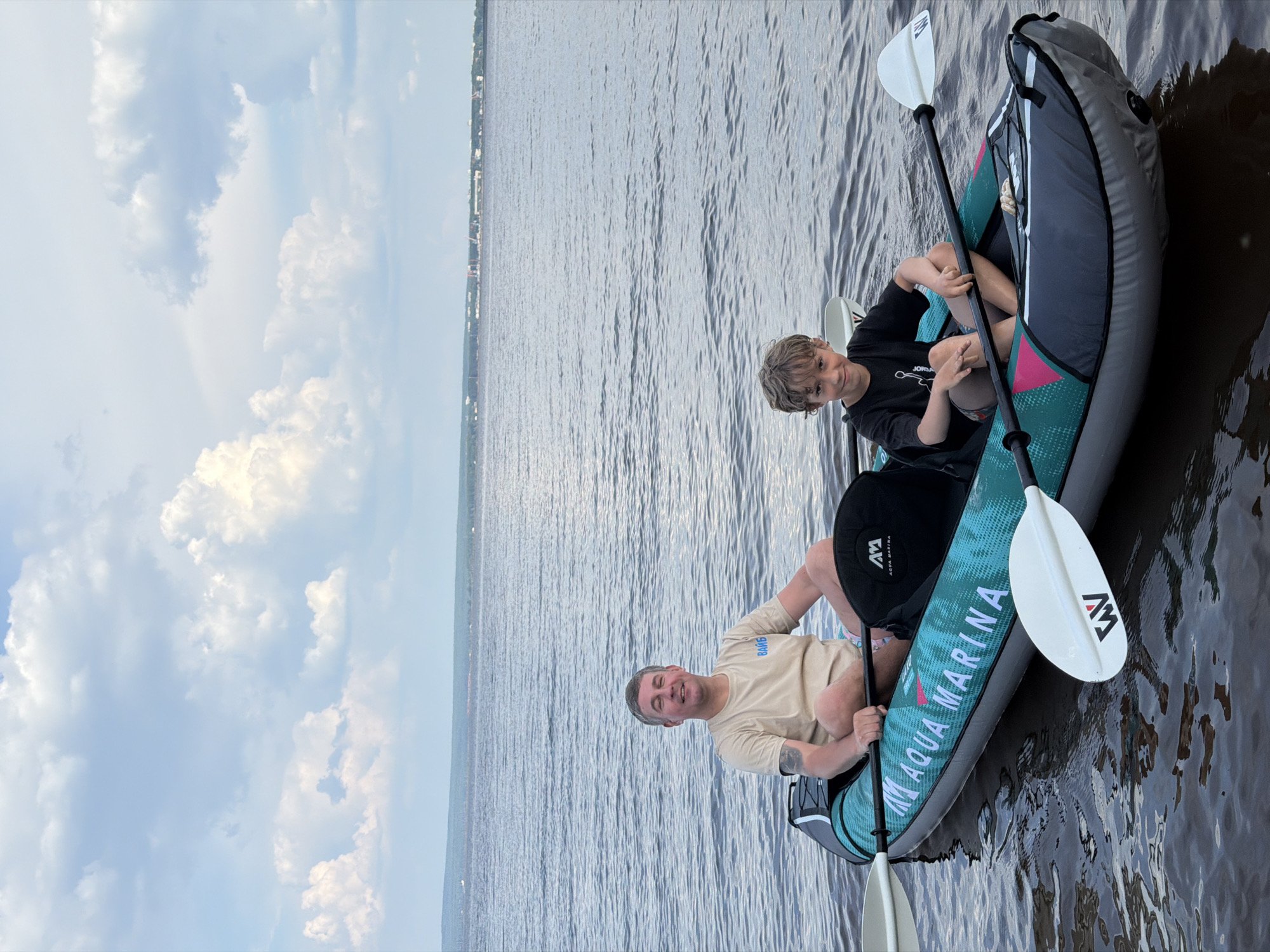 An outdoor, eye-level, medium shot captures two individuals, a man and a boy, seated in an inflatable two-person kayak on a body of water. The man, positioned in the front of the kayak, faces forward and smiles at the camera. He has short gray hair, a light complexion, and wears a light tan t-shirt with the word "ВАЙБ" on the back. His left arm is bent, and his hand rests on the paddle. The boy, located in the rear of the kayak, also faces forward and looks directly at the camera with a slight smile. He has light brown, curly hair and wears a black t-shirt with a graphic design. His hands are clasped in his lap. Both individuals are wearing casual clothing suitable for outdoor activities.
The kayak is dark gray and teal with "AQUA MARINA" written in white lettering along its side. It is partially submerged in the water, which appears calm with small ripples. Two white paddles with black handles are visible, one on each side of the kayak, with their blades resting in the water. The background consists of a vast body of water that stretches to a distant, hazy shoreline. A faint line of trees and structures can be seen along the horizon, suggesting a populated area. The sky above is filled with a mixture of white and gray clouds, with patches of blue sky visible. The overall lighting suggests it is daytime, likely late afternoon or early evening, given the soft light. The image appears to have been taken from a slightly elevated perspective, looking down at the kayak. The scene implies a recreational outing on a river or lake, possibly near the city of Noviy, Russia, though this cannot be definitively confirmed from the visual information alone.