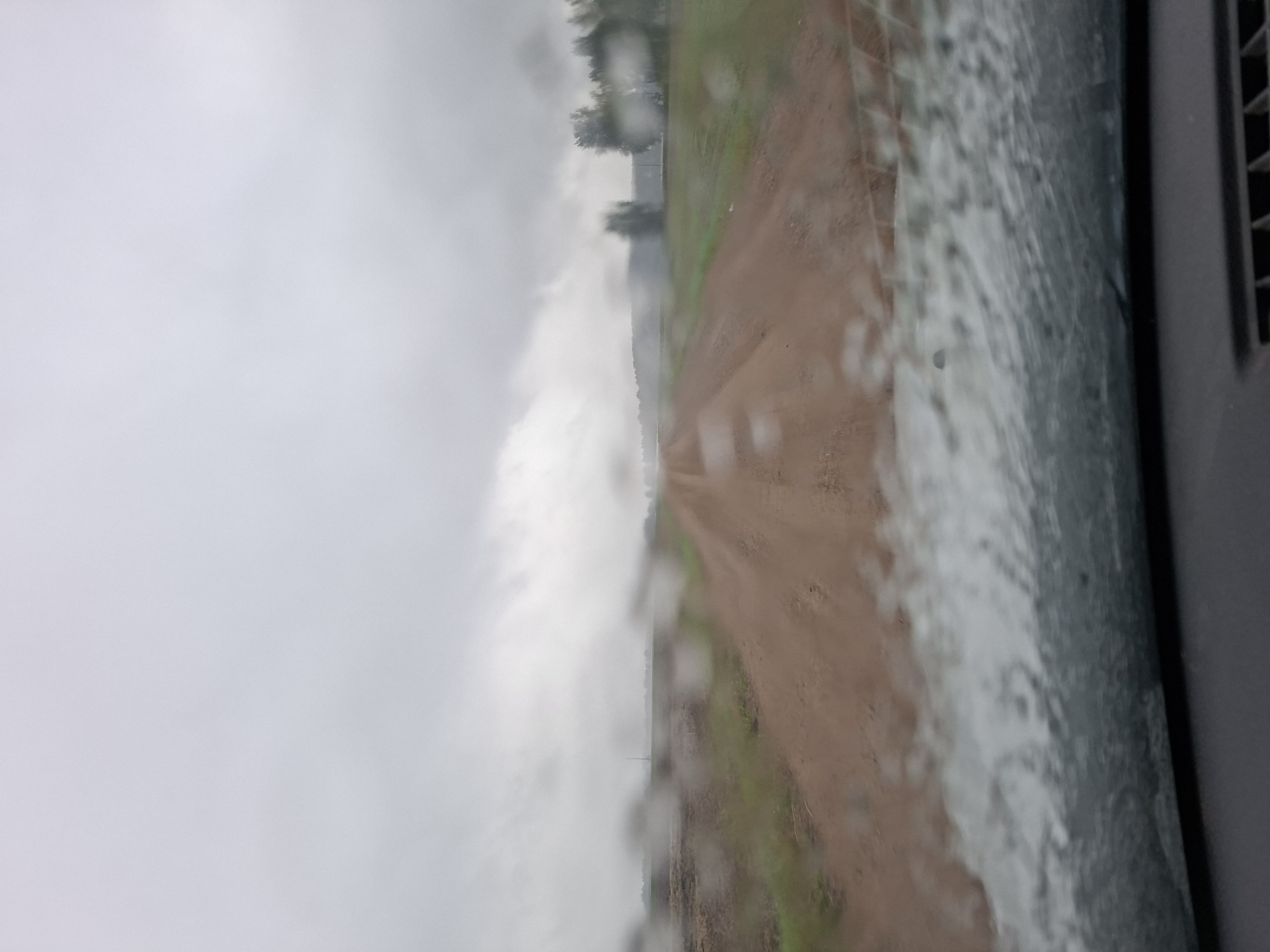 The image depicts a view from inside a vehicle looking out through a rain-streaked windshield onto a dirt road. The road appears to be unpaved and is surrounded by green grass and some trees in the distance. The sky is overcast and grey, suggesting it is raining or has recently rained. The perspective is looking forward down the road, which curves slightly to the right in the distance. The foreground shows the interior of the car, with the dashboard visible at the bottom. There are no people or animals visible in the scene. The location is identified as Славенскі сельскі Савет, Belarus.
