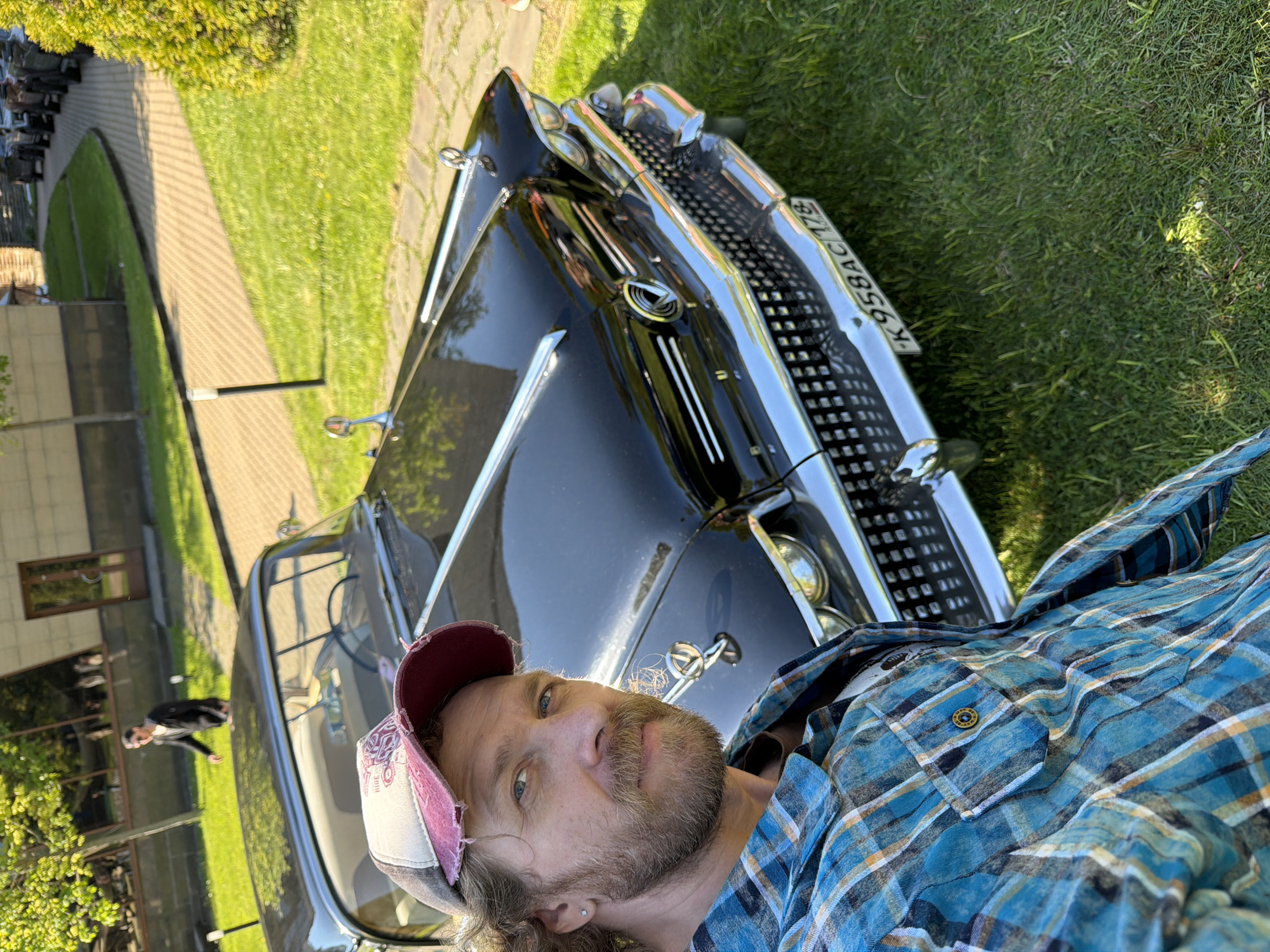 A man is taking a selfie with a black vintage car, likely a GAZ-13 Chaika, parked on a grassy area. The car's front grill and license plate "K958AC178" are visible. In the background, there is a building, a paved walkway, and more greenery. The man is wearing a blue plaid shirt and a distressed baseball cap. The overall location is Repino, Russia. A second man is visible near the building in the background.