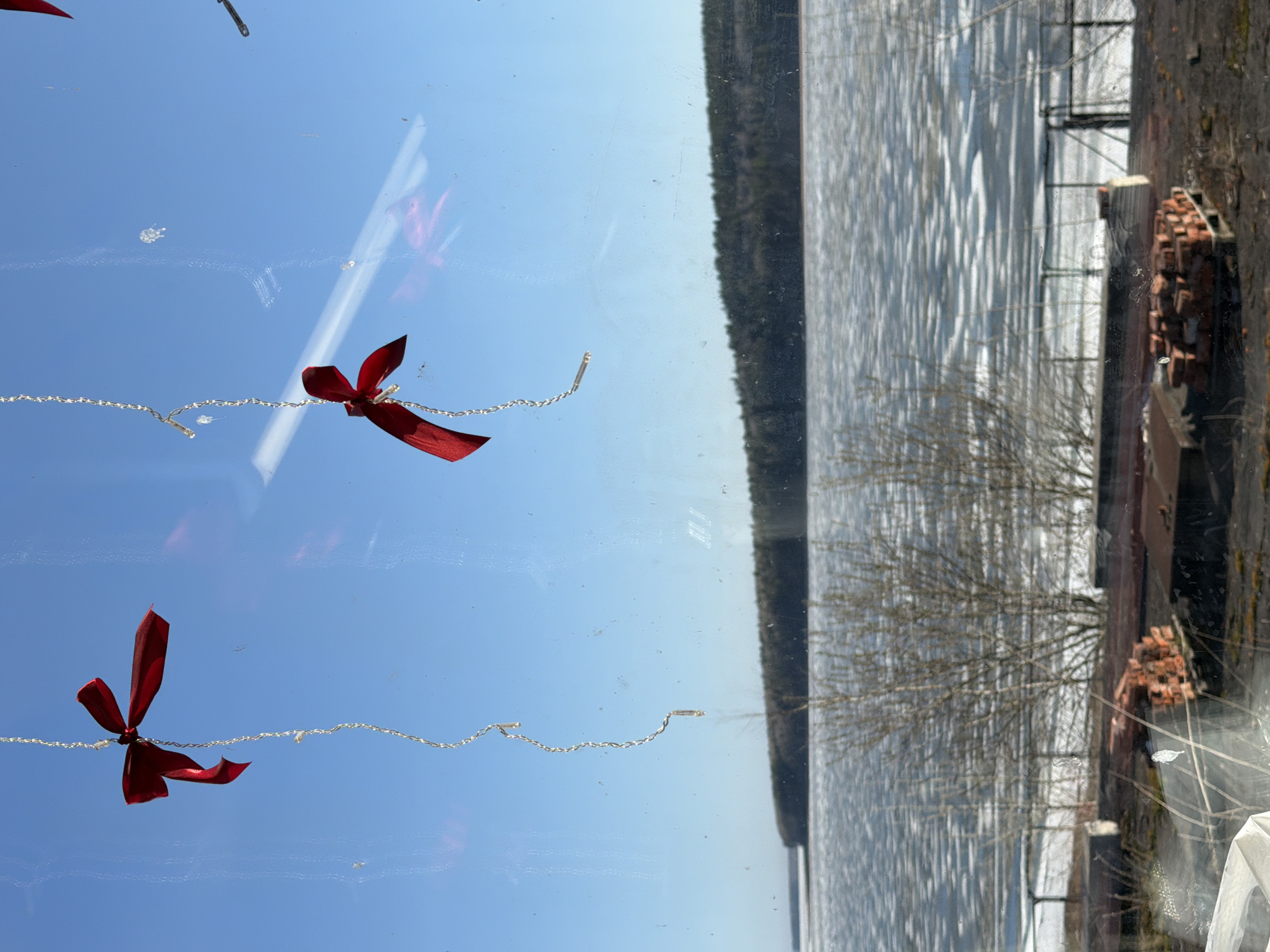 The image shows a view from a window. Two red bows with attached silver string are taped to the inside of the window. Outside, there is a partially frozen body of water, likely a river or lake, with a dark treeline along the far shore. On the right side of the frame, there is a brick structure, possibly a building or wall, with a pile of bricks. Bare tree branches are visible in front of the structure. The sky is blue and clear, reflected in the window. The location is Chaikovsky, Russia.