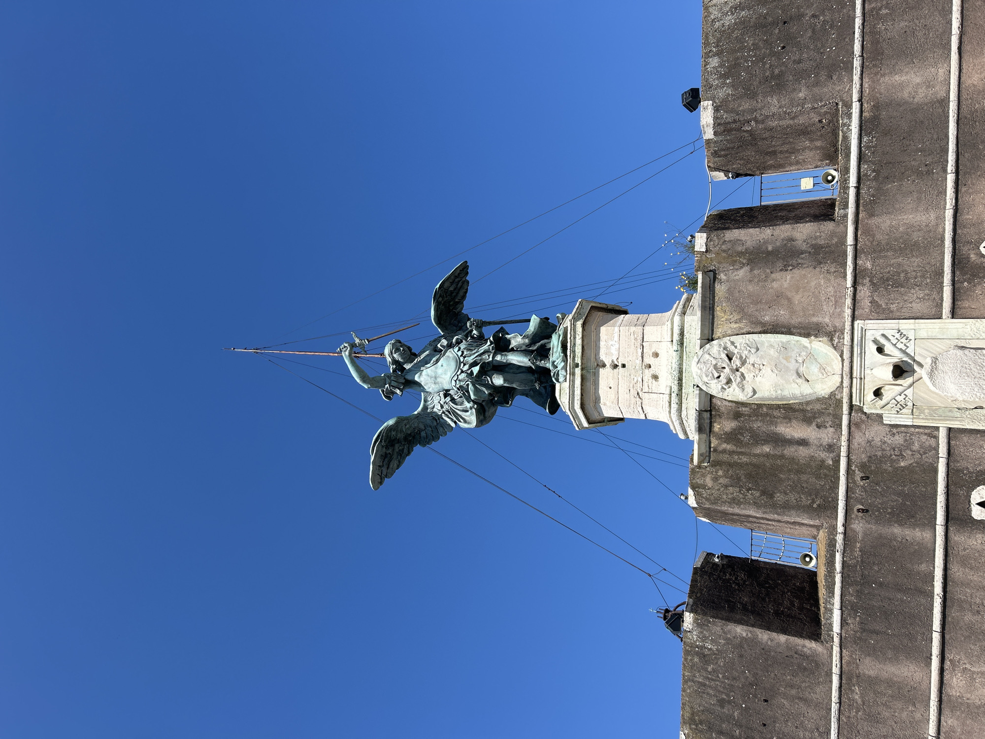 The image displays a bronze statue of Archangel Michael situated atop the Castel Sant'Angelo in Rome, Italy. The winged figure wears armor and drapery, with its right arm raised, holding a sword pointed skyward. The sword's blade integrates into a reddish-brown, vertical mast, possibly a lightning rod or antenna. The statue rests on a multi-tiered, light-colored stone pedestal featuring carved reliefs and a coat of arms. Below the pedestal are the dark grey, weathered stone battlements of the fortress, showing signs of age and minor plant growth. A metal railing is visible behind the statue's base. On the left battlement, a dark, angular device, possibly a security camera or light, is mounted. A similar device is on the right battlement. Within an embrasure on the right battlement, a metal gate is visible, along with a white horn-shaped speaker and a small white sign. Multiple thin, dark wires extend from the statue's upper structure and base, connecting to points on the battlements. The background is a clear, unclouded blue sky. No human figures are present. The perspective is a low-angle upward view.