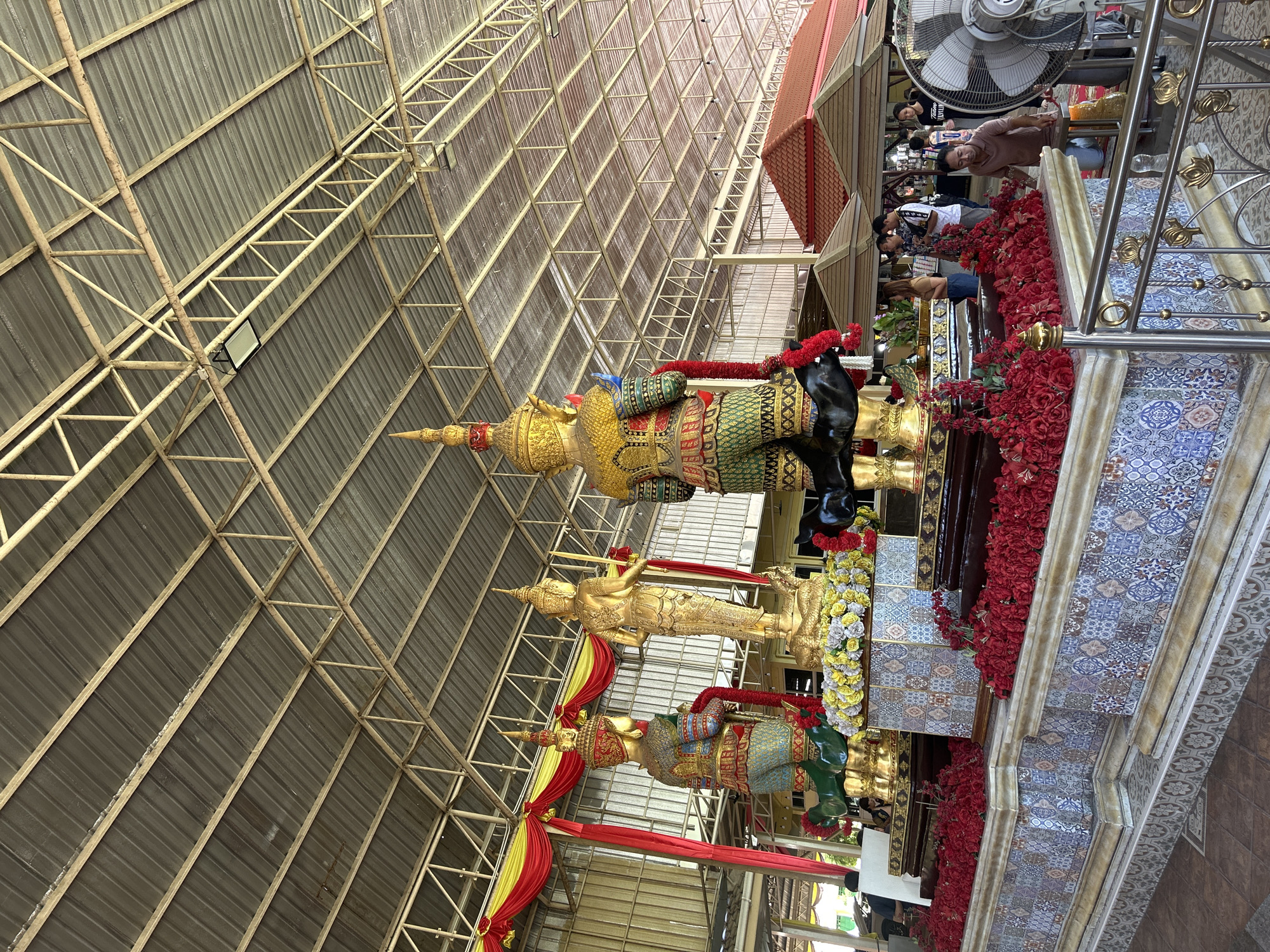 The image shows a covered outdoor area with a corrugated metal roof supported by a metal frame structure. In the foreground, there are three large, ornate statues. The statue on the left is a standing figure in green and gold attire with a tall headdress, holding a red garland. The central statue is a golden figure with multiple arms, standing on a coiled serpent. To the right, another large statue, similar in design to the one on the left, is partially visible. All statues are adorned with red and yellow garlands and flowers. The base of the statues is decorated with colorful mosaic tiles and surrounded by arrangements of red roses.
In the background, several people are visible, some standing and observing the statues, while others appear to be walking or engaged in activities. A large industrial fan is positioned to the right of the frame. The overall environment suggests a temple or a cultural site, possibly open to visitors. The presence of the Thai Ko Subdistrict, Thailand, indicates the geographical location of this scene.