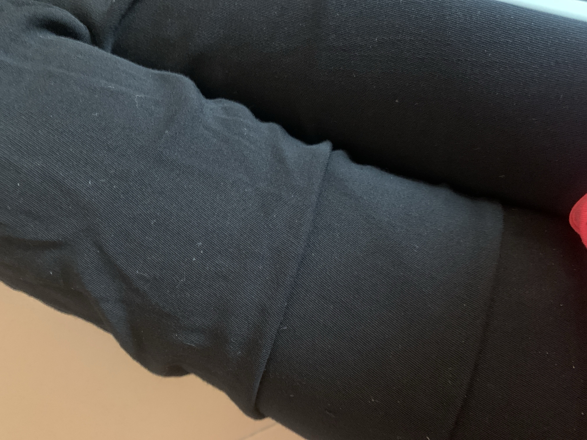 The image is a close-up, angled shot of black fabric, likely clothing, with visible texture and some lint. The fabric appears to be folded or bunched, creating shadows and highlights that accentuate the ribbed texture. The background is a blurred, light brown color. There is no information available about this image being located in Jauro Yinu, Nigeria.
